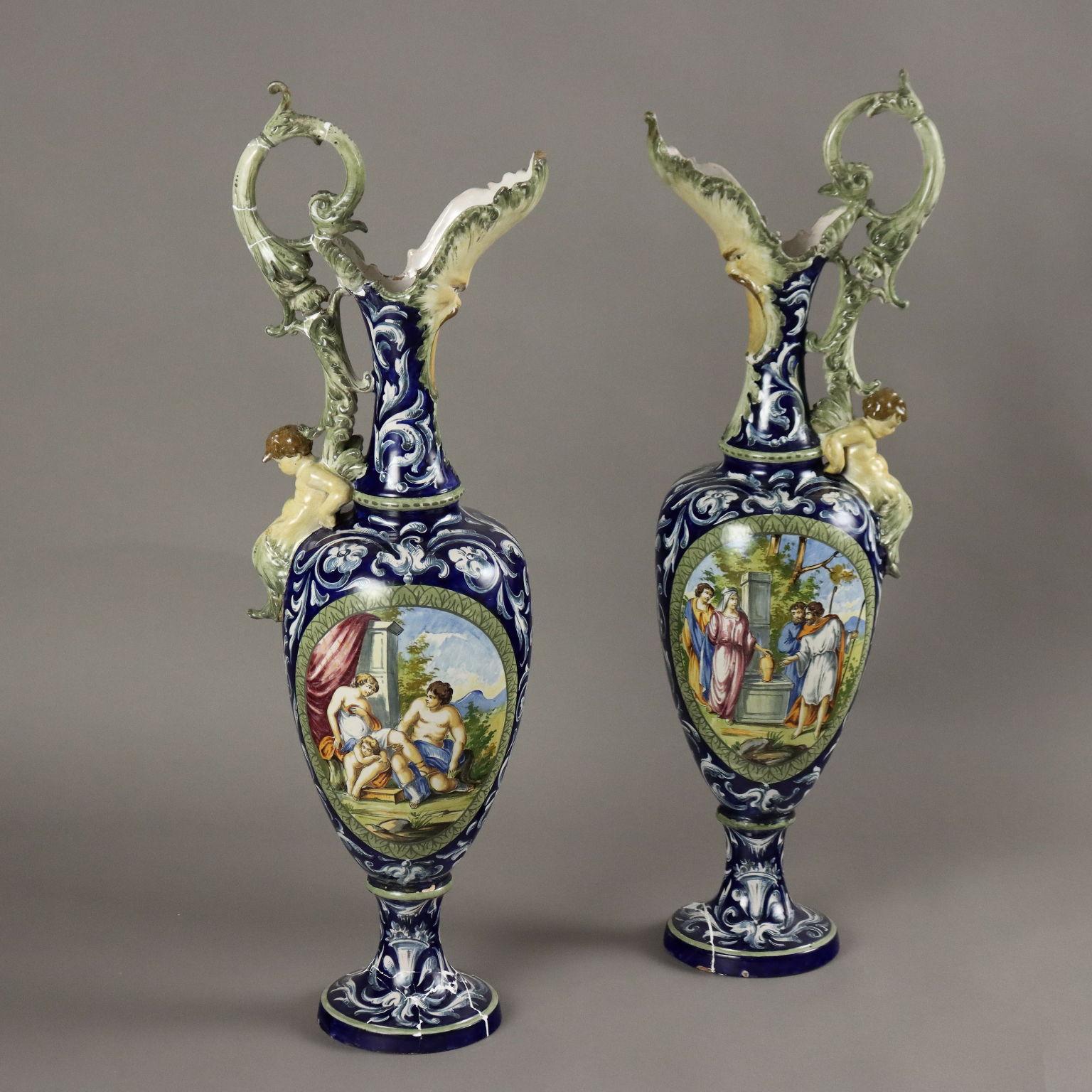 Paar glasierte Steingutamphoren, verziert mit Pflanzenmotiven, Masken und geflügelten Drachen auf blauem Grund. Die Amphoren weisen Malereien mit Figuren und Putten in den Reserven, einen Ausguss mit einer großen Szenenmaske sowie eine Blattvolute
