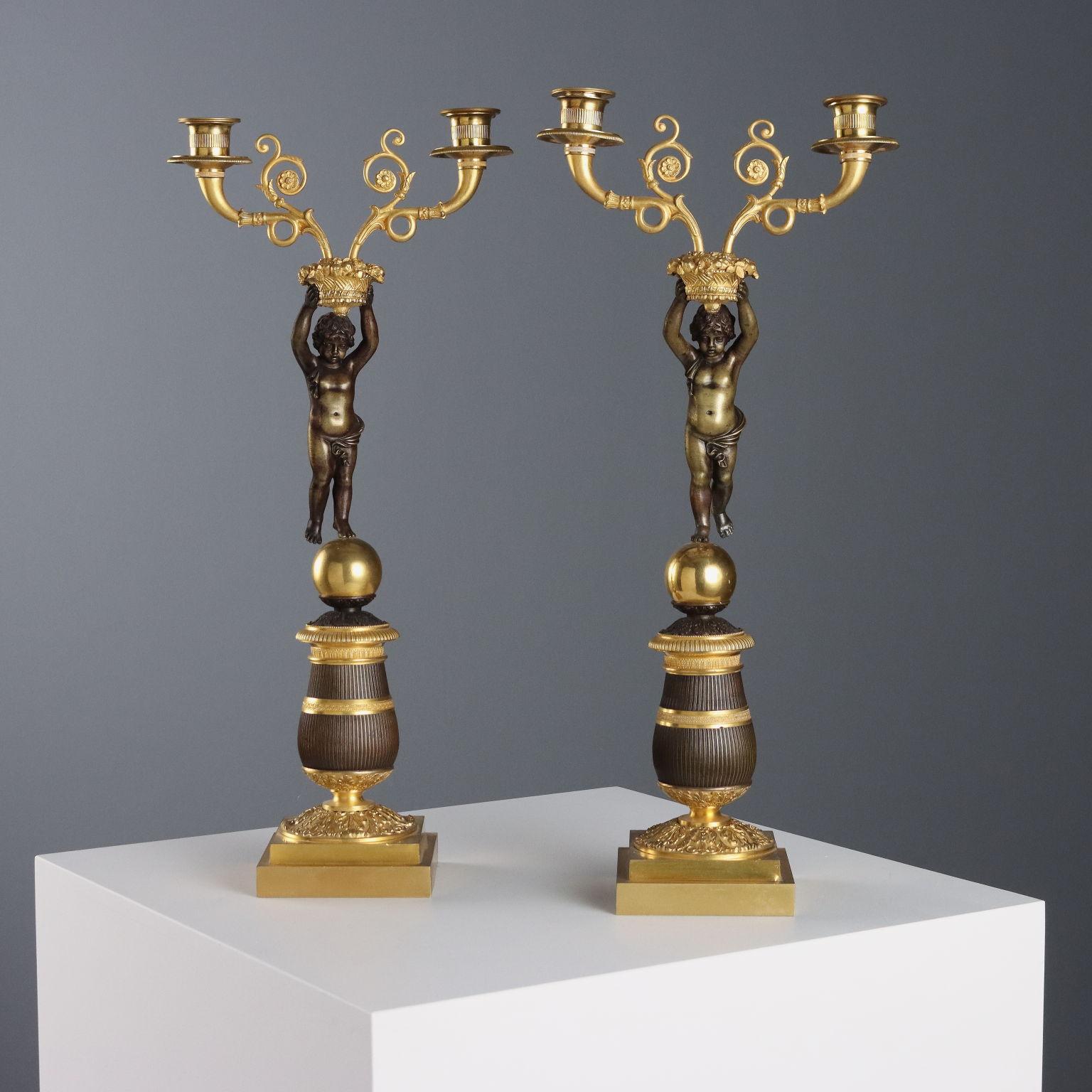 Coppia di candelabri a due luci in bronzo dorato e cesellato. Bracci a volute fogliacee poggianti su cesto di frutta retto da putto in bronzo a patina scura. Il putto poggia su sfera e plinto a forma di vaso decorato a scanalature e da elementi