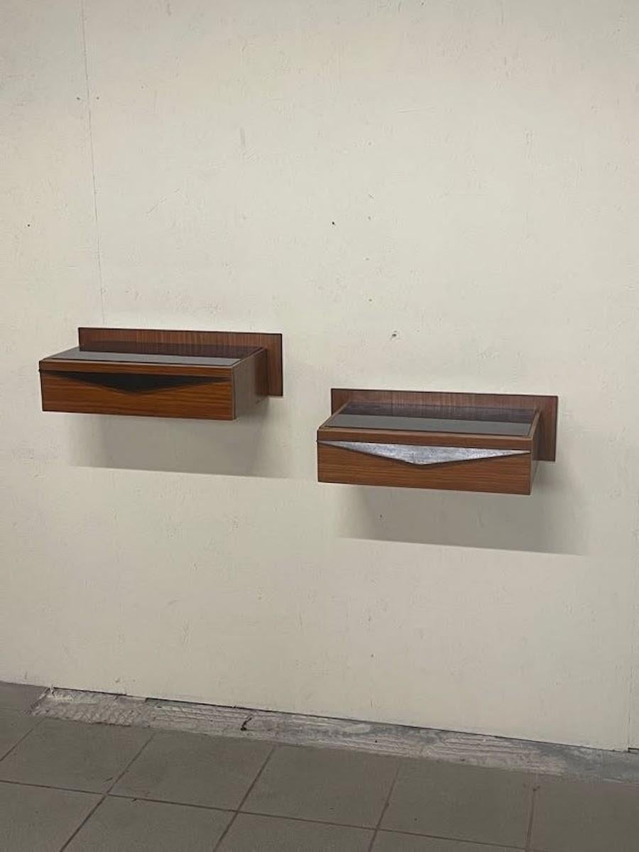 Coppia di comodini a parete anni '60.

Struttura in pregiato legno di mogano e parti in legno ebanizzato.
Top in vetro blu originale.

Ciascun comodino è corredato di un pratico cassetto.

Completamente restaurati.

Sono perfetti con una lampada a