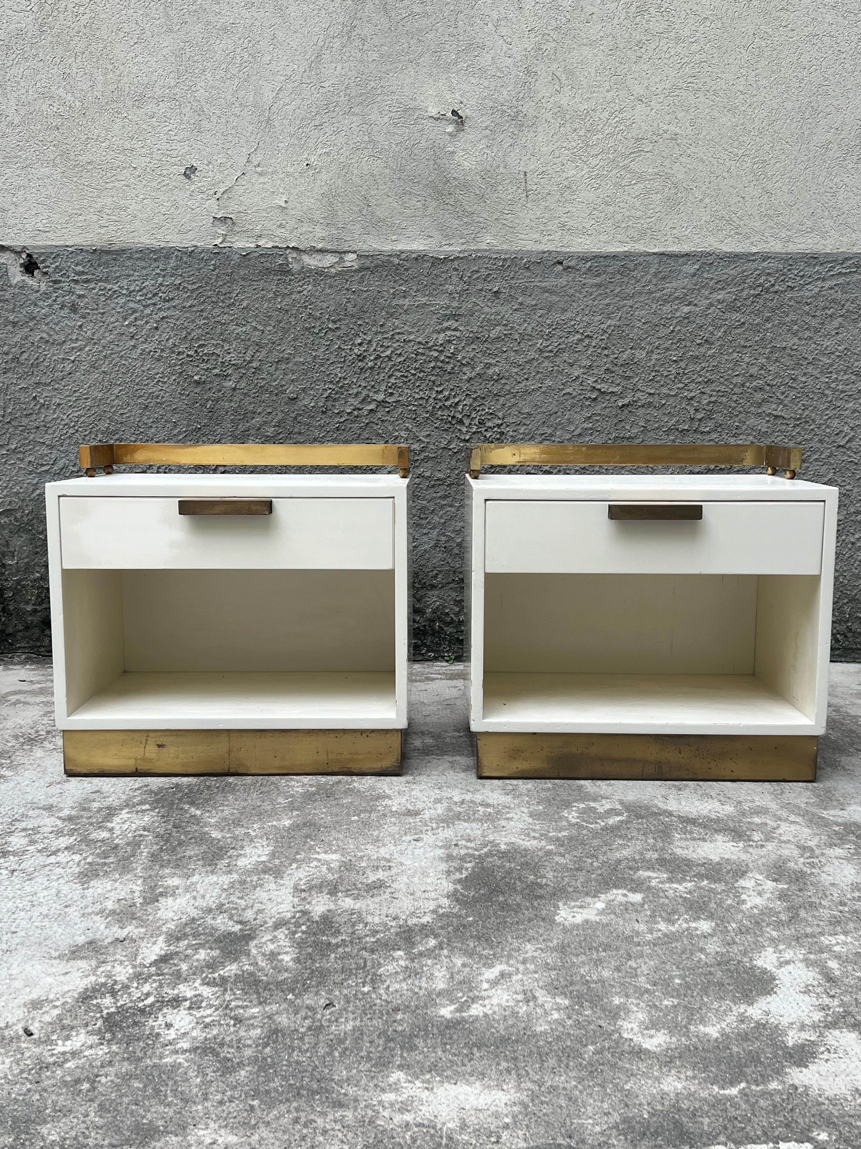 Elegant pair of bedside tables in ivory lacquered wood, enhanced by brass details such as a beading to cover the perimeter band of the base, a convenient, solid handle in the center of the drawers, and a functional balustrade that rests on spheres,