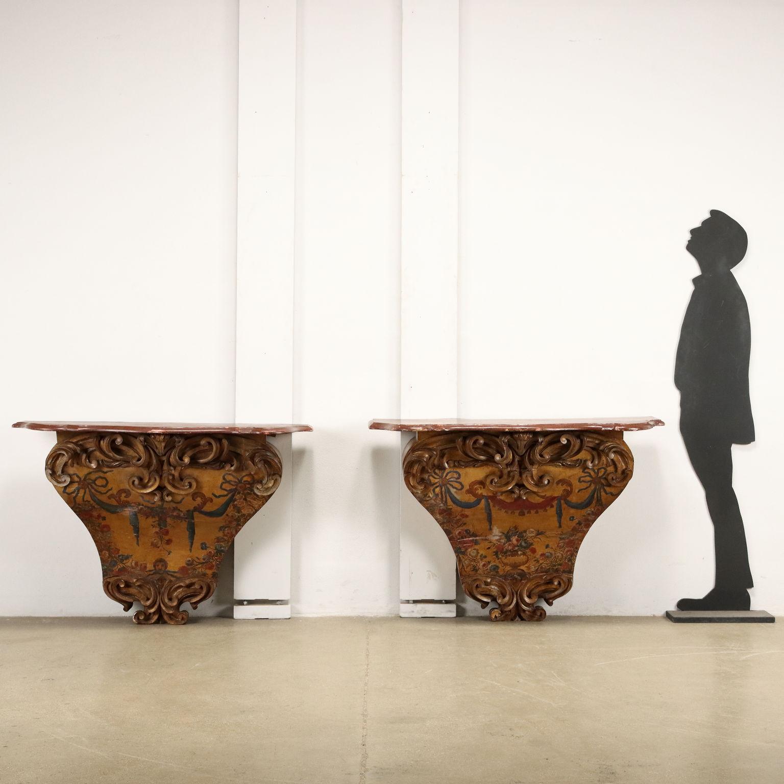 Coppia di consolle in legno laccato e dorato, impreziosite con intagli a ricciolo nel sottopiano e alla base, corpo centrale e fianchi dipinti a motivi neoclassici con festoni  e decorazioni floreali. Piano in legno in finto porfido rosso.
Sud