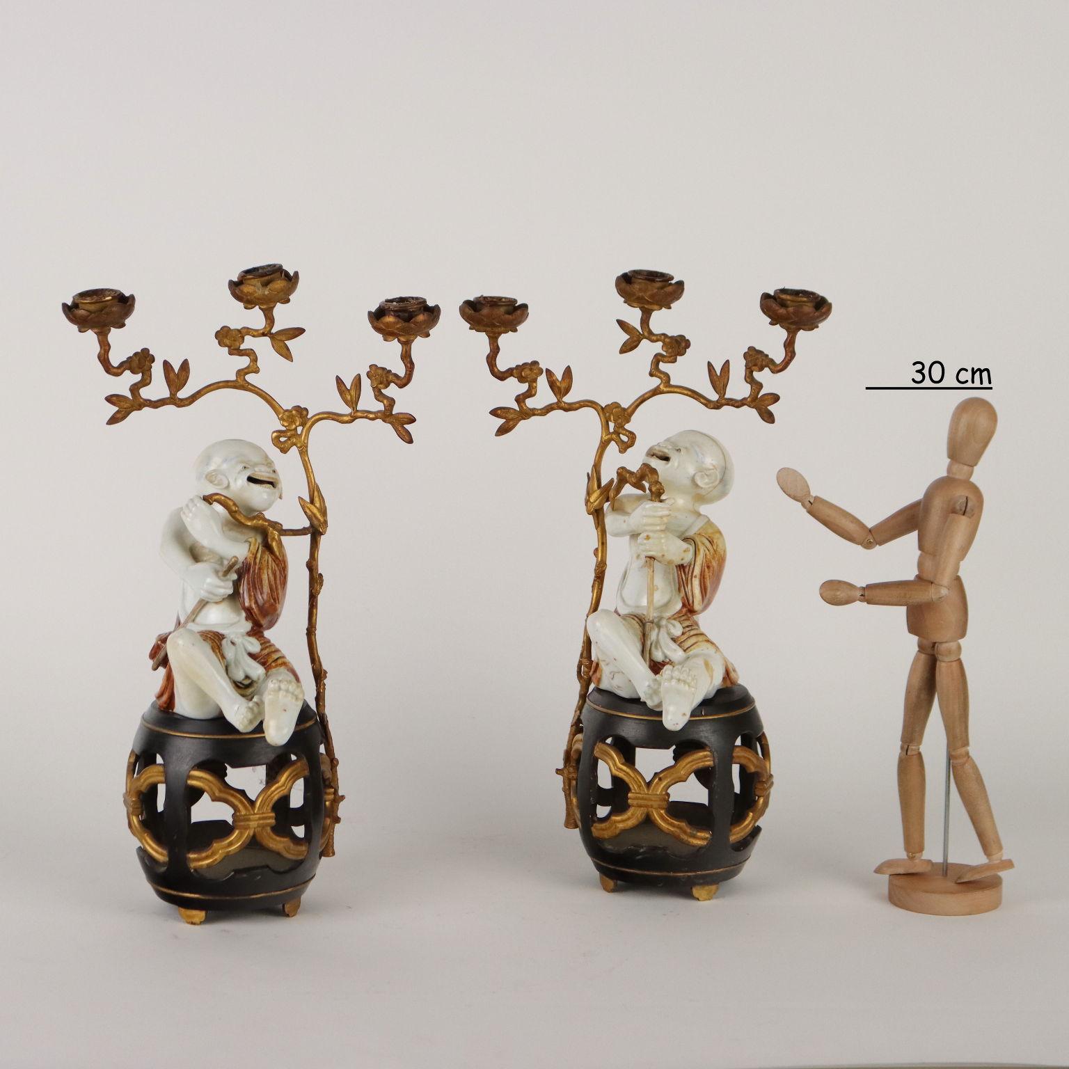 Coppia di raffinati portacandele con soggetti evocativi in porcellana montati su sedili da giardino in bronzo brunito e dorato. I due fratelli 