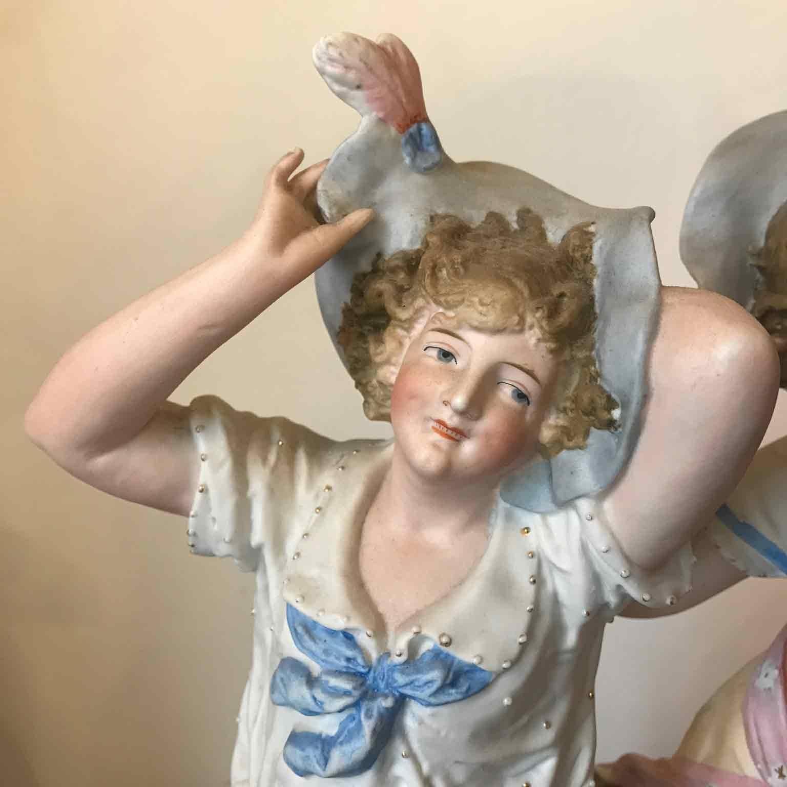 Coppia di Sculture italiane in Porcellana Biscuit raffiguranti due figure femminili con le vesti al vento,  due giovani ragazze intente a trattenere  il cappello, in una giornata ventosa, su basamenti quadrangolari. Queste statue in biscuit sono