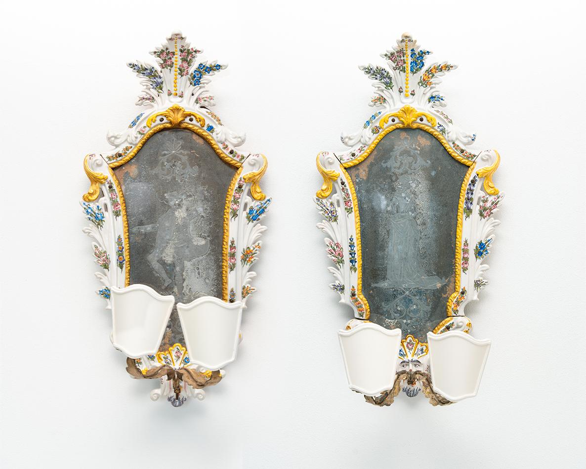 Questa bellissima coppia di specchierine in maiolica furono realizzate in Veneto nel XVIII secolo.

Hanno cornici sagomate in maiolica bianca con fiori arricchiti da dorature.
Alla base è presente un mascherone dalla cui bocca si dipartono due