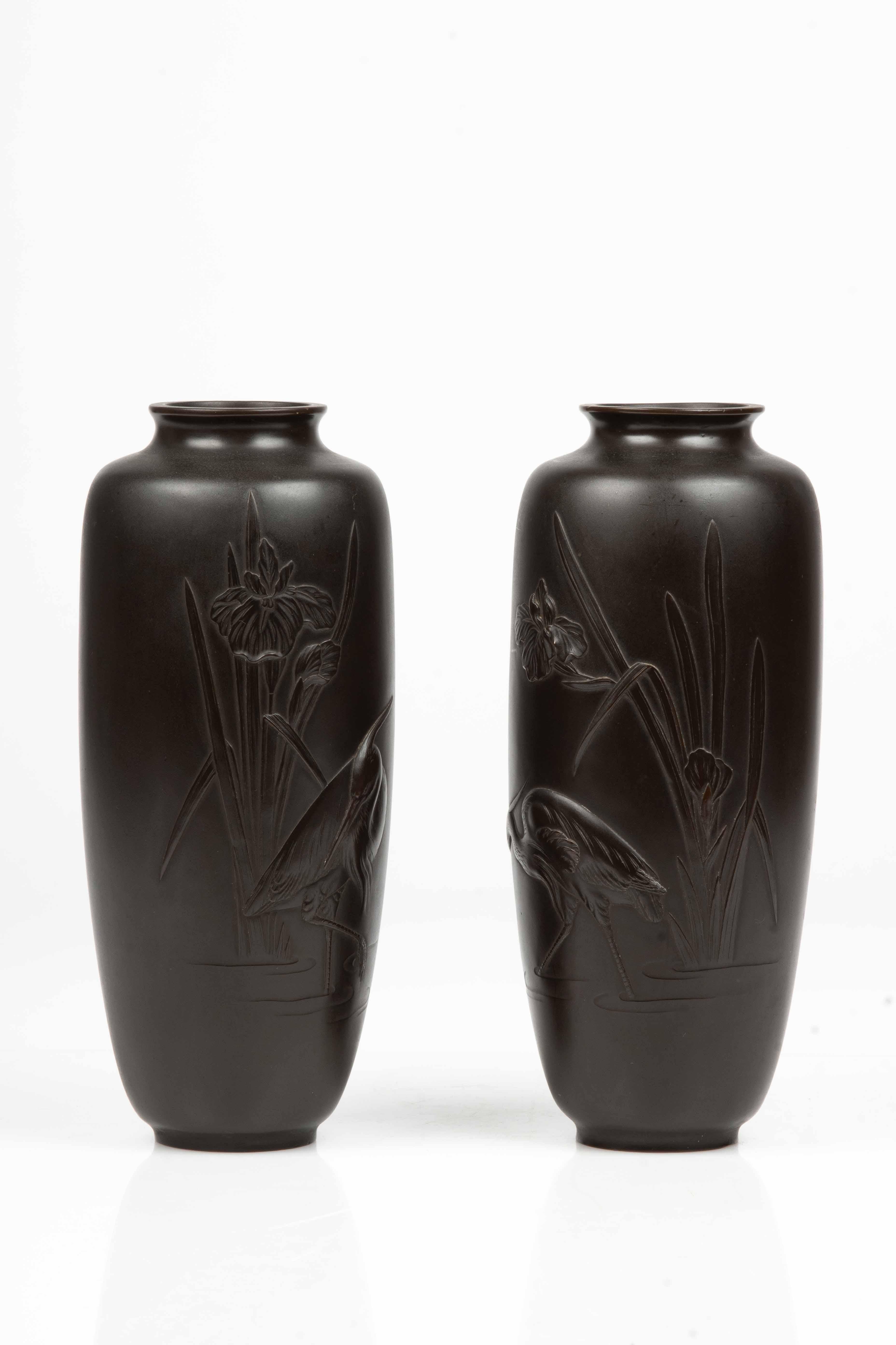 Una coppia di vasi in bronzo giapponesi dalla forma cilindrica e patina marrone scuro, raffiguranti una coppia di garzette vicino a un ruscello, con le zampe immerse nell’acqua e circondate da due piante di iris in fiore. Firma Masayuki è incisa