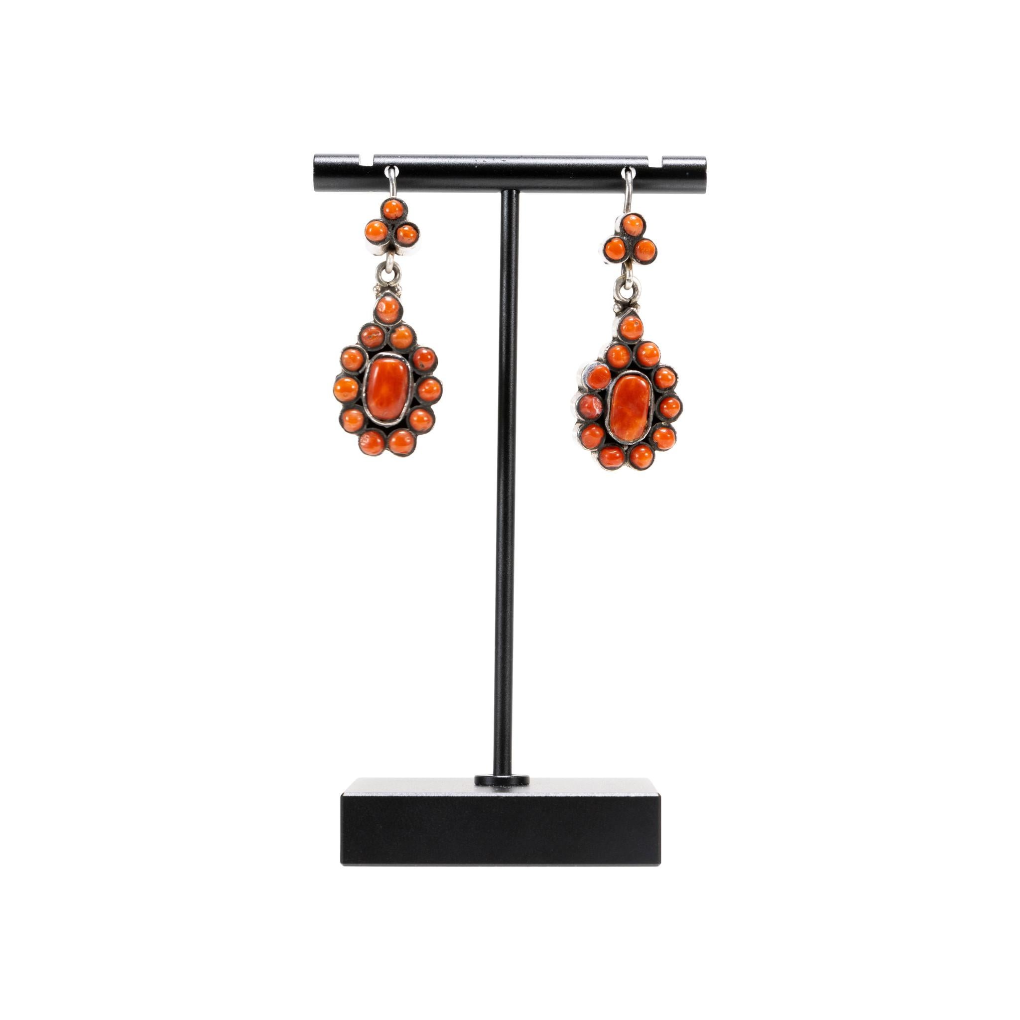 Boucles d'oreilles pendantes en corail et argent. Le corail est d'une belle couleur orange brûlé et est disposé en grappe avec un cabochon ovale plus grand au centre. Entourées de 11 petites perles de corail, elles sont attachées à un lien avec