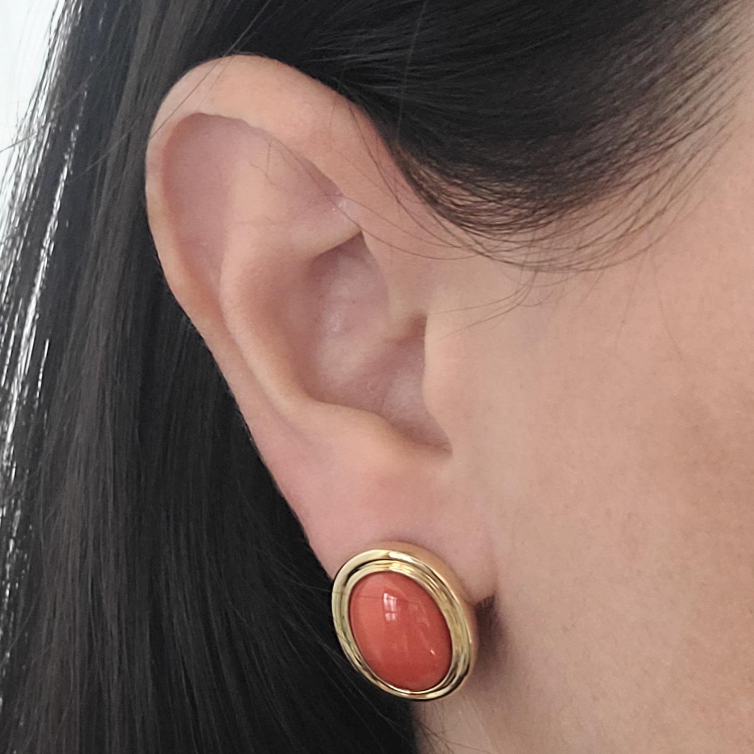 14 Karat Yellow Gold Classic Coral Stud Earrings. These Timeless Clip-On Studs 2 Bezel Set Oval Cabochon-cut Bright Orangey-Red Corals Measuring Approximately 15mm x 11mm. Pierced Post with Supportive Hinged Omega Back; Post Removed Upon Request.
