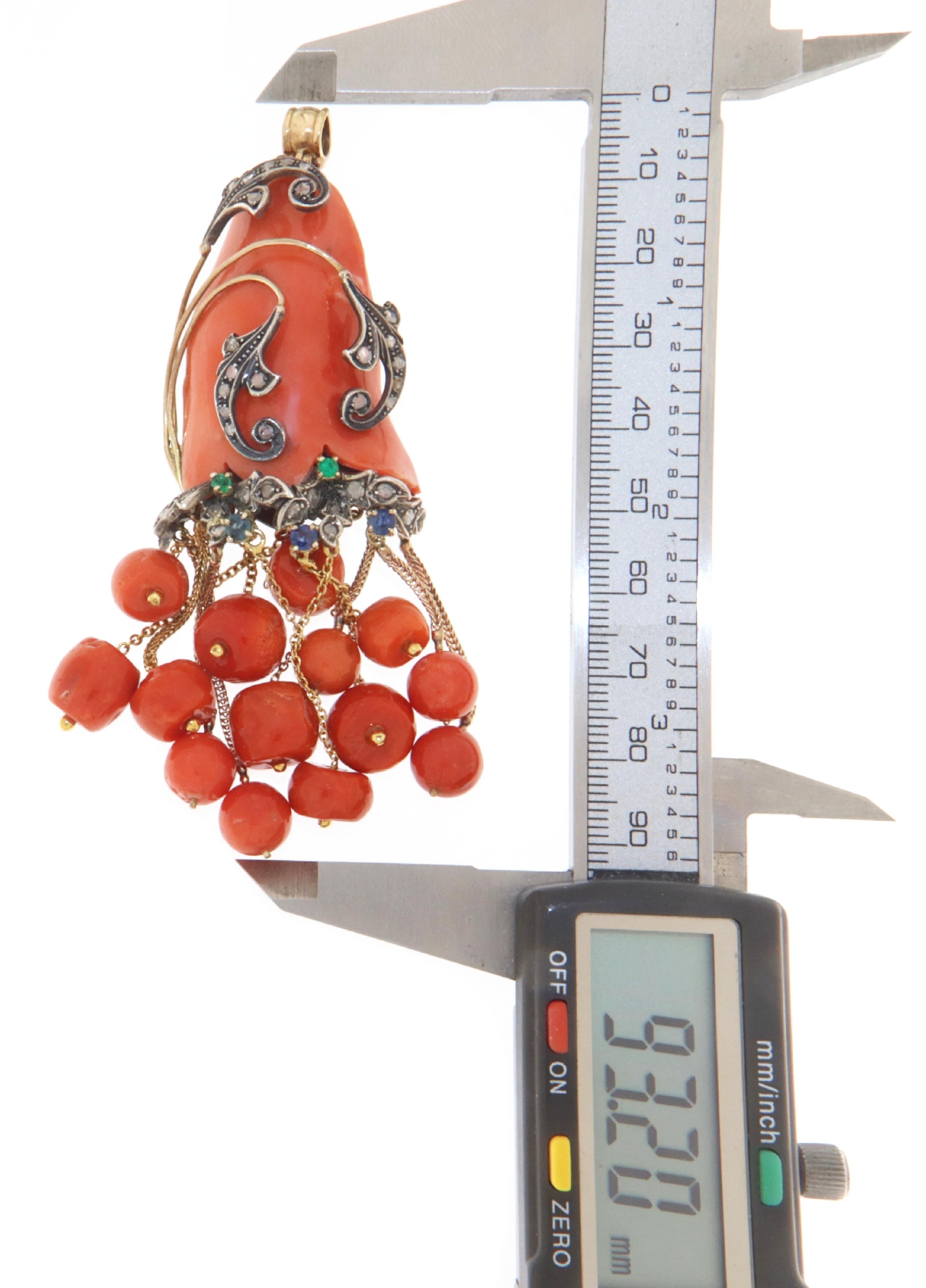 Spectacular retro pendant made entirely by hand in 18-karat yellow gold and 800 thousandths silver.
An irregularly shaped orange coral is enriched by flourishes on which are set antique rose cut diamonds and emeralds and sapphires that accentuate