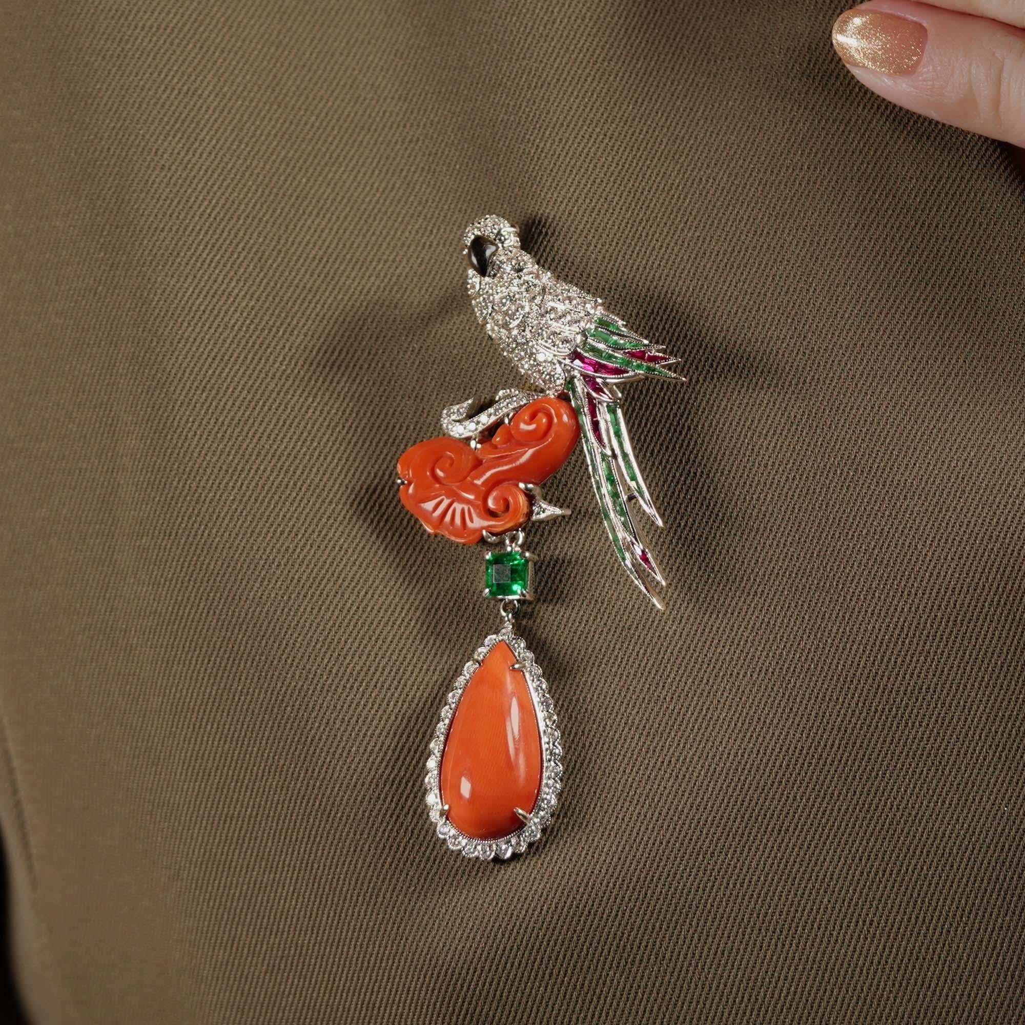 Vogelbrosche im Art-Déco-Stil mit einem mit Diamanten besetzten Vogel auf einer Korallenwolke. Smaragde formen die gefiederten Flügel des Vogels und verleihen ihm lebendige Eleganz. Ein Smaragd im Quadratschliff verbindet das Nest mit einem