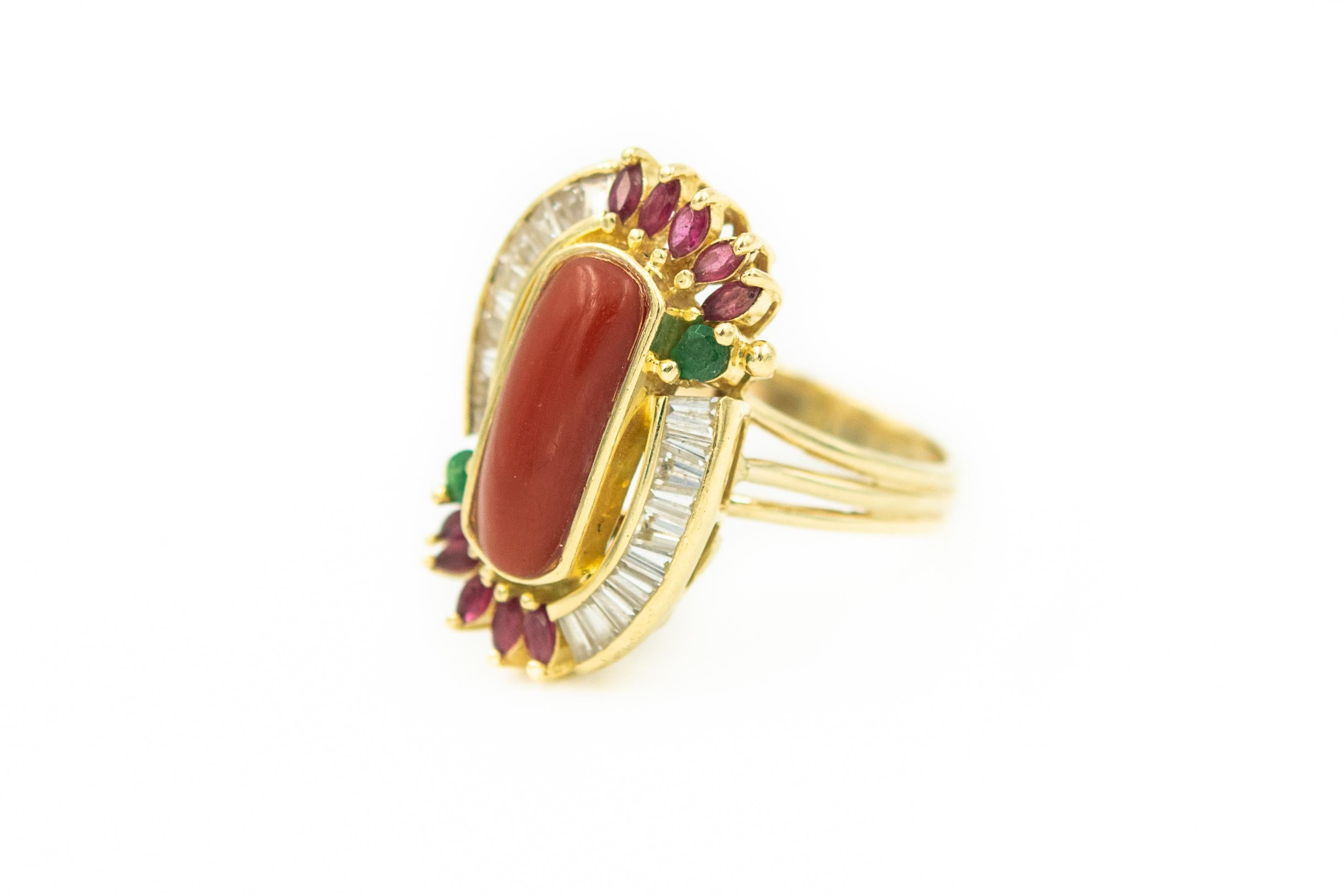 Late 20th Century cocktail ring featuring a lozenge shaped bezel set coral slab accented by 10 prong set marquise shaped rubies, 2 round emerald and 20 white sapphire baguette shaped stones.  The stones are mounted in a 14k yellow gold ring.  The