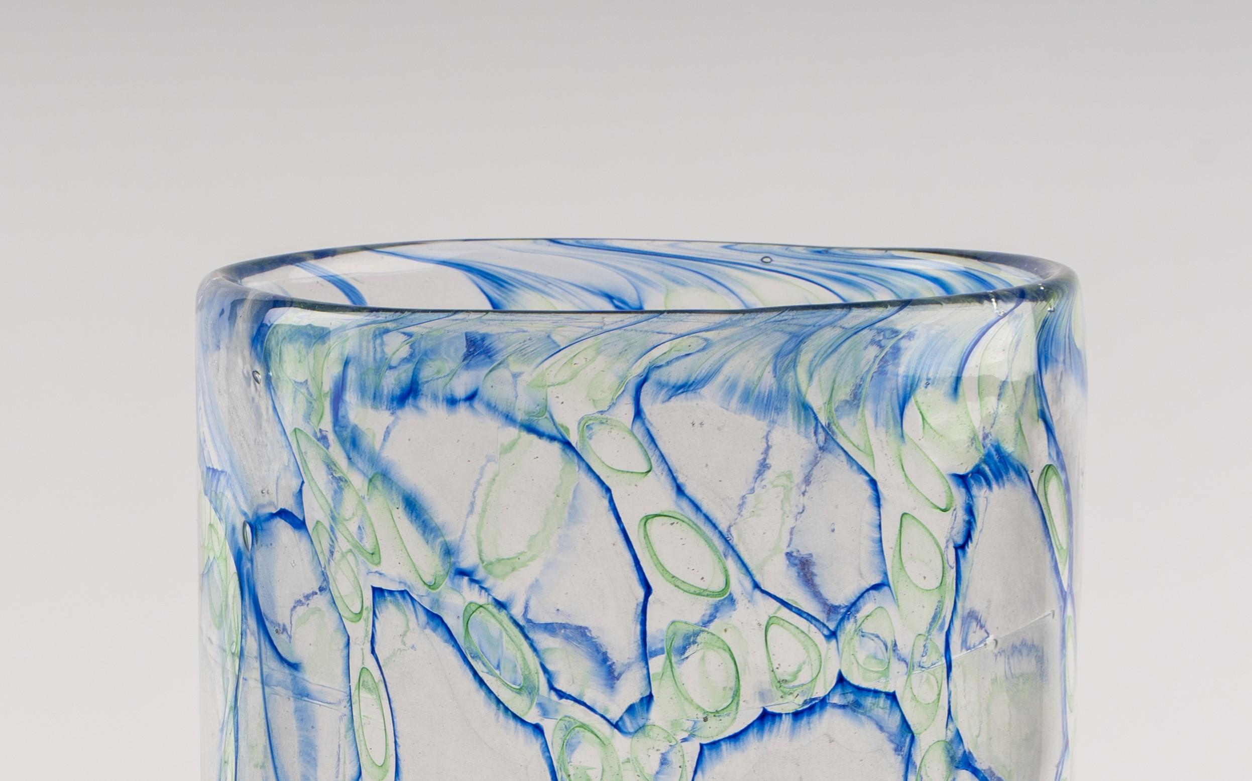 Jeremiah Jacobs, a masterful glassblower, has created an exquisite blown glass piece that incorporates the captivating murrini technique. This stunning artwork features a coral-like pattern formed by blue and green lines on clear glass, showcasing