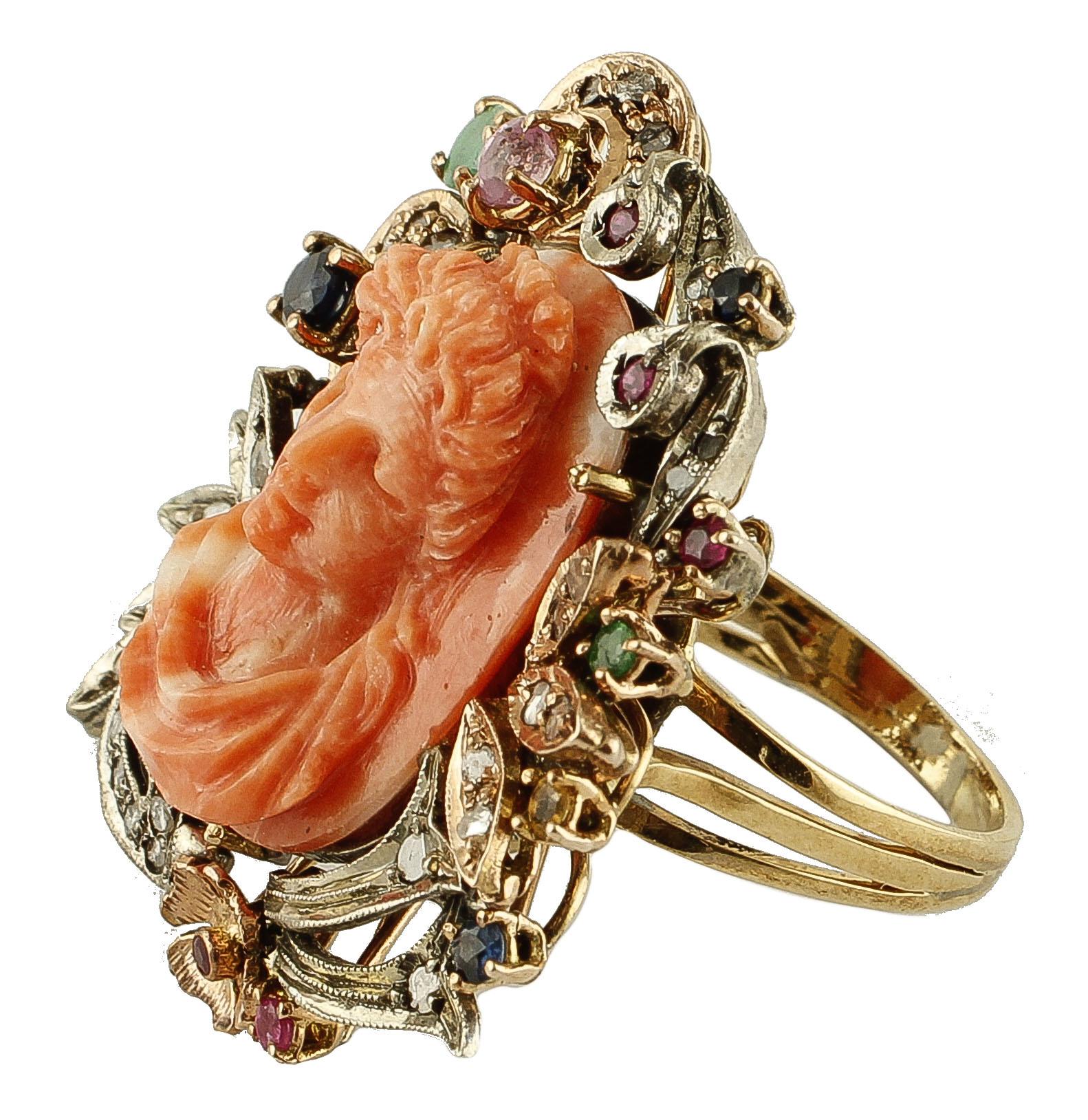 Beautiful retro ring in 9k rose gold and silver structure, mounted with a central elatius coral finely carved with lady motif, framed by rose gold and silver flowery decorations studded with diamonds, emeralds, rubies and blue sapphires. 
This ring