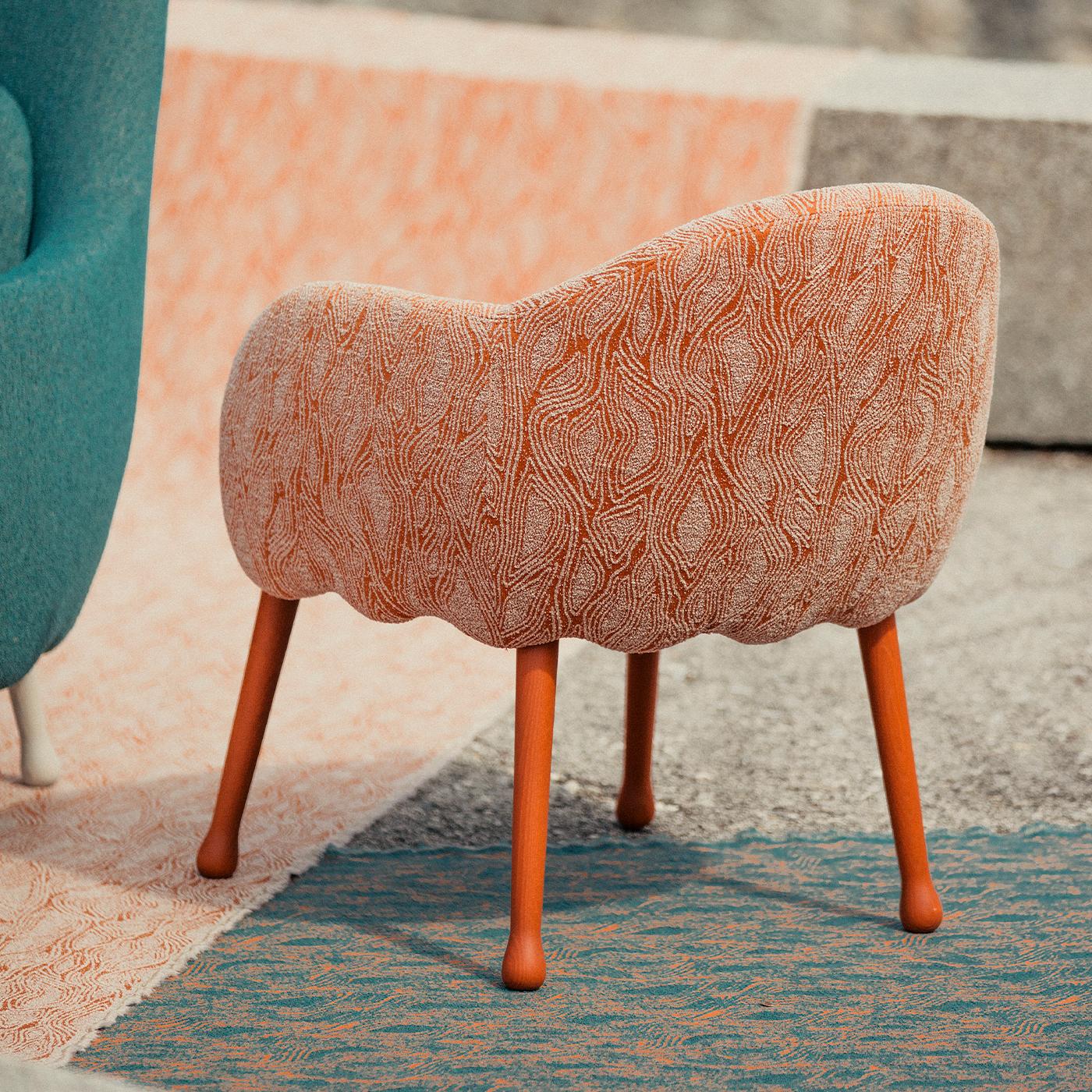 Ce fauteuil glamour imprégnera les intérieurs modernes d'un charme éclectique, souligné par l'inspiration florale révélée par le bord festonné qui souligne le profil inférieur de l'assise et par les pieds en forme de tige, laqués orange, en hêtre