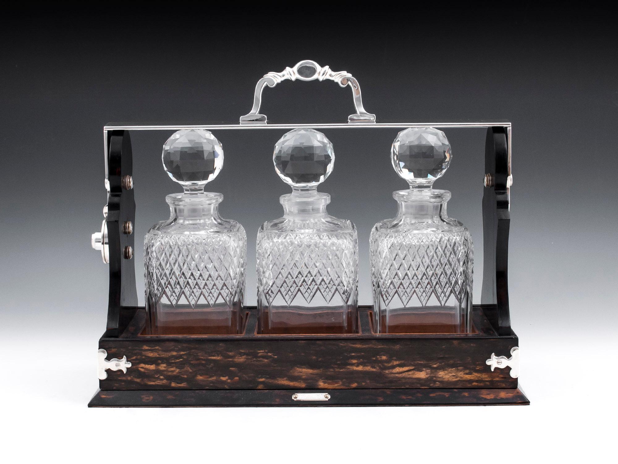 Betjemann Tantalus with silver plate handle and corner brackets, veneered in stunning Coromandel retailed by Mappin Brothers, London. 

The antique tantalus has three diamond shaped crystal cut glass decanters with faceted stoppers. Complete with