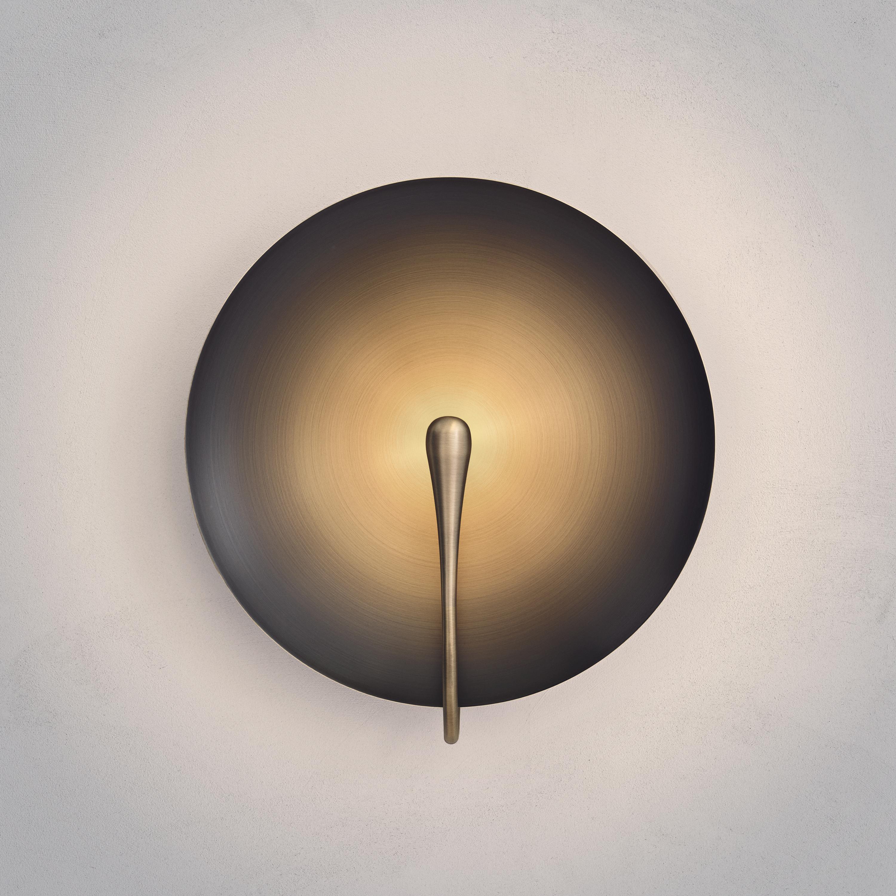 A gentle silhouette of a plate makes up this unique wall light. To create this orbital, gradient-finished shade, a mixed patina is applied on a hand-spun brass plate, from dark bronze to bright brushed brass, to accentuate the light effect. Please