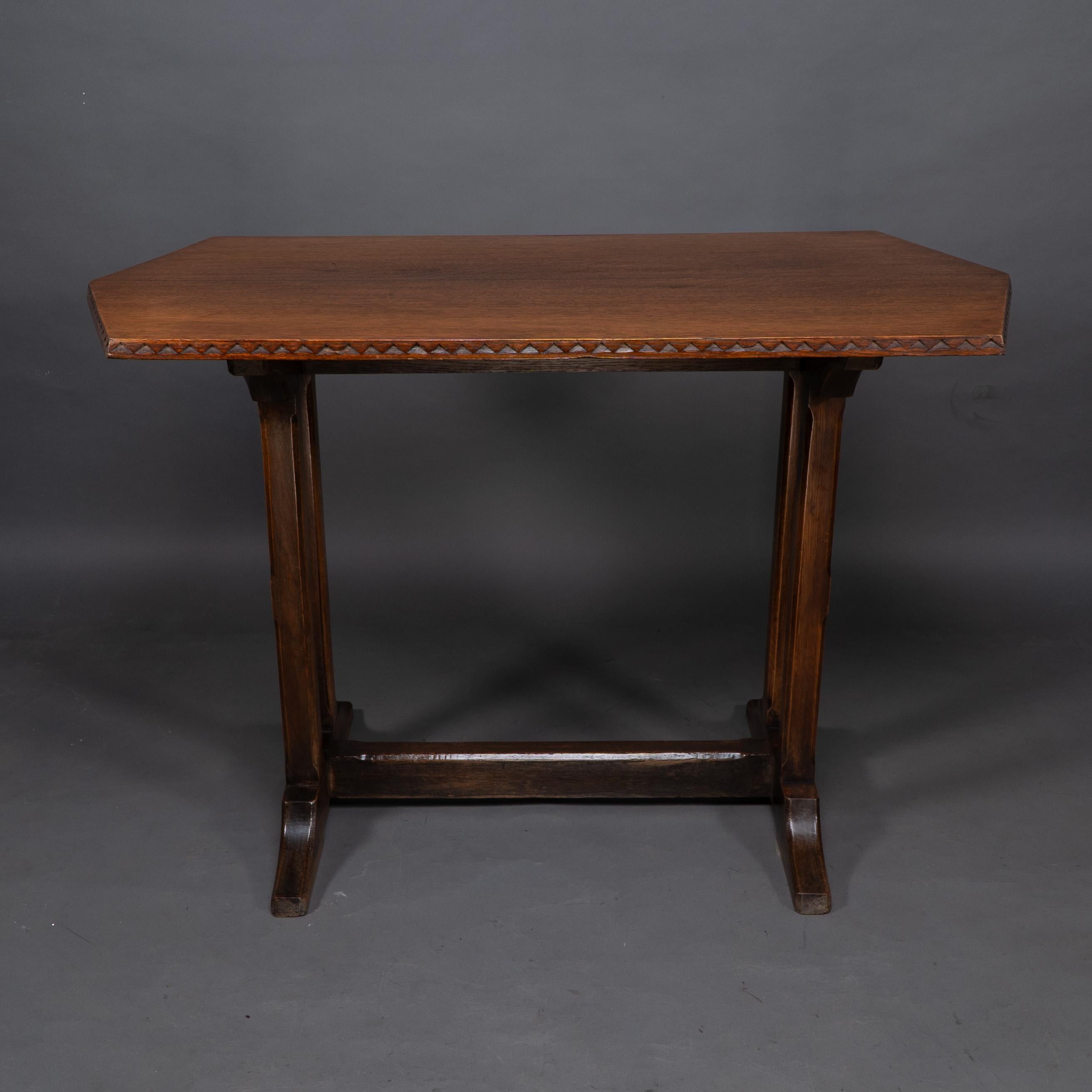 Table de réfectoire ou de bibliothèque en chêne de style Cotswold, avec des chevrons sculptés sur les bords du plateau à double angle. Sur deux pieds droits aux arêtes chanfreinées à 45 degrés qui s'unissent aux pieds en forme, l'entretoise