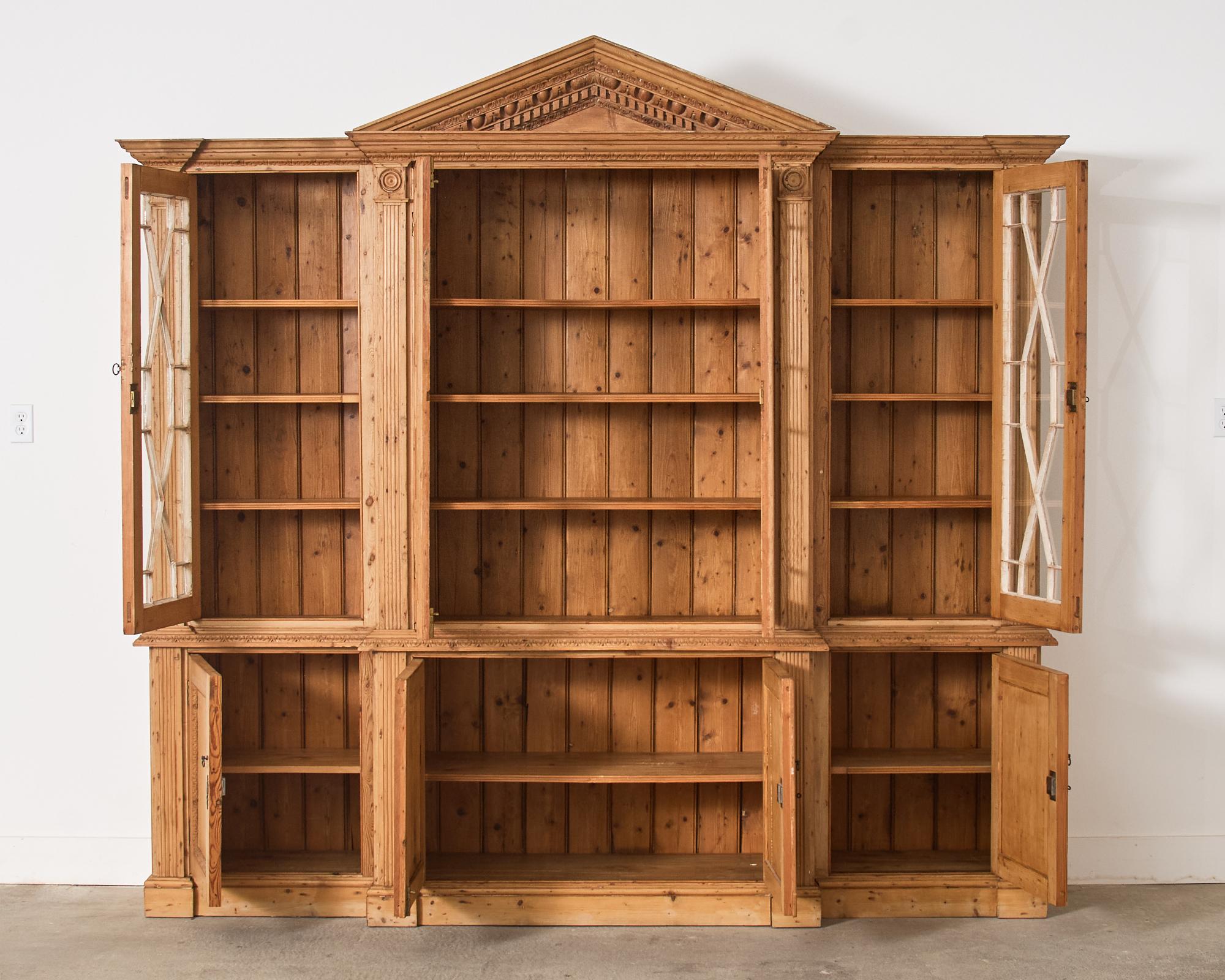 Grand late 19th/early 20th century country English library breakfront bookcase cabinet crafted from pine. Made in the Georgian taste with intricate carved neoclassical motifs. The top of the case features four astragal glazed windows. The four