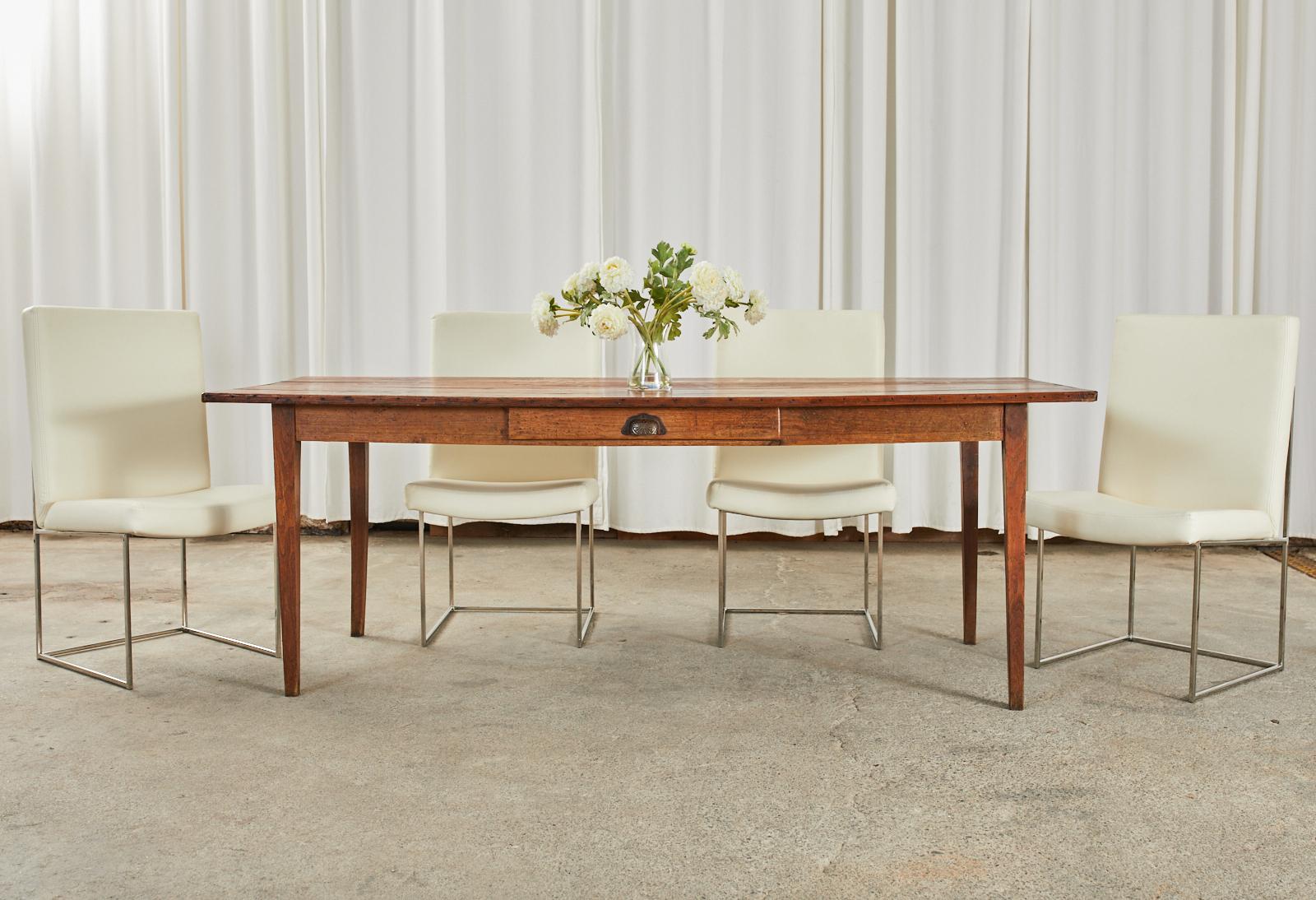 Elegant country English farmhouse dining or harvest table crafted from pine. The farm table features a 4 plank top with a thin border trim. There is a storage drawer in the frieze on one side with a patinated brass pull. Constructed with wood peg