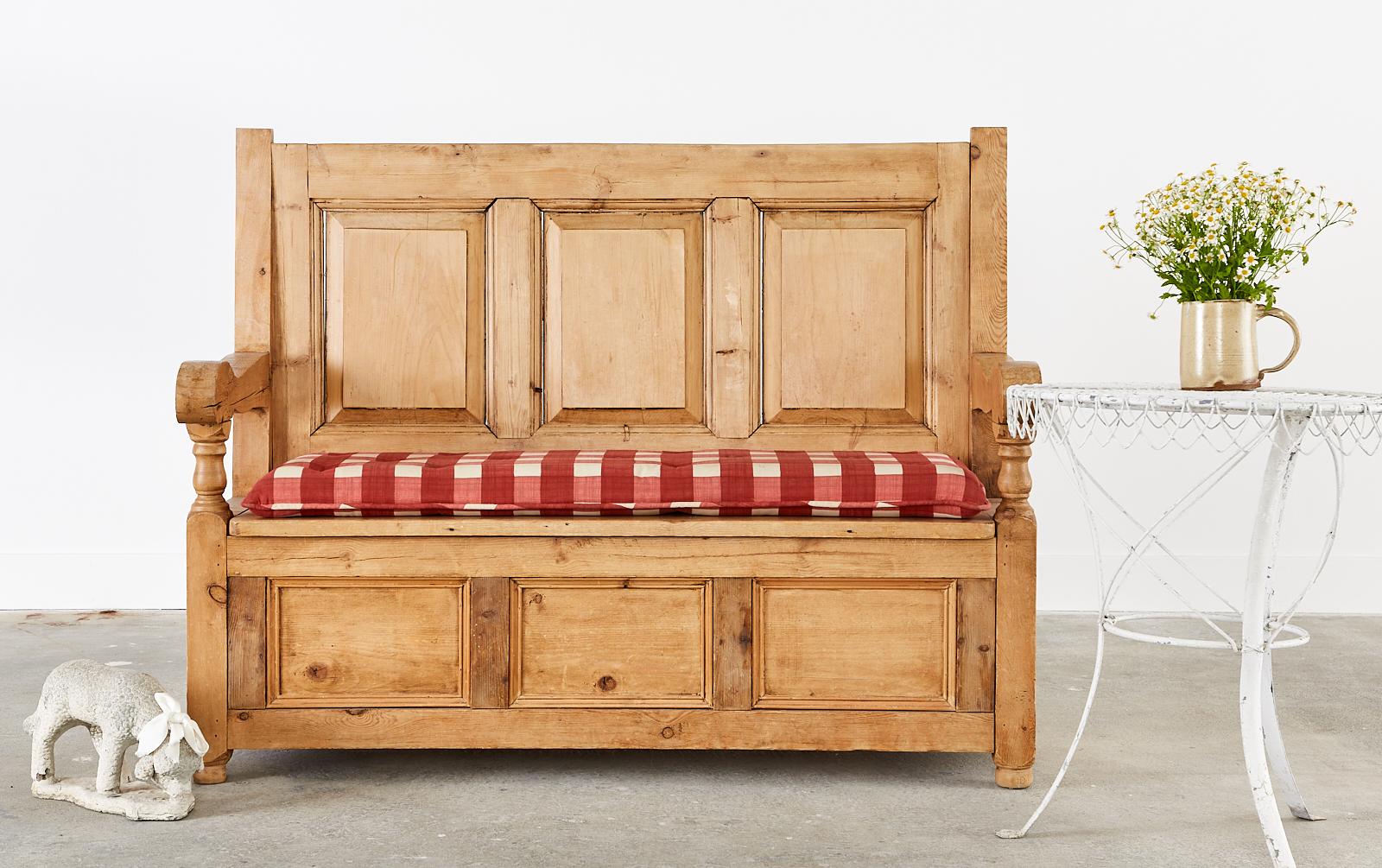 Charming and rustic country English provincial farmhouse settle bench seat or settee crafted from pine. The bench is constructed with peg joinery and has decorative panels on three sides. The arms and legs have thick, chunky proportions measuring