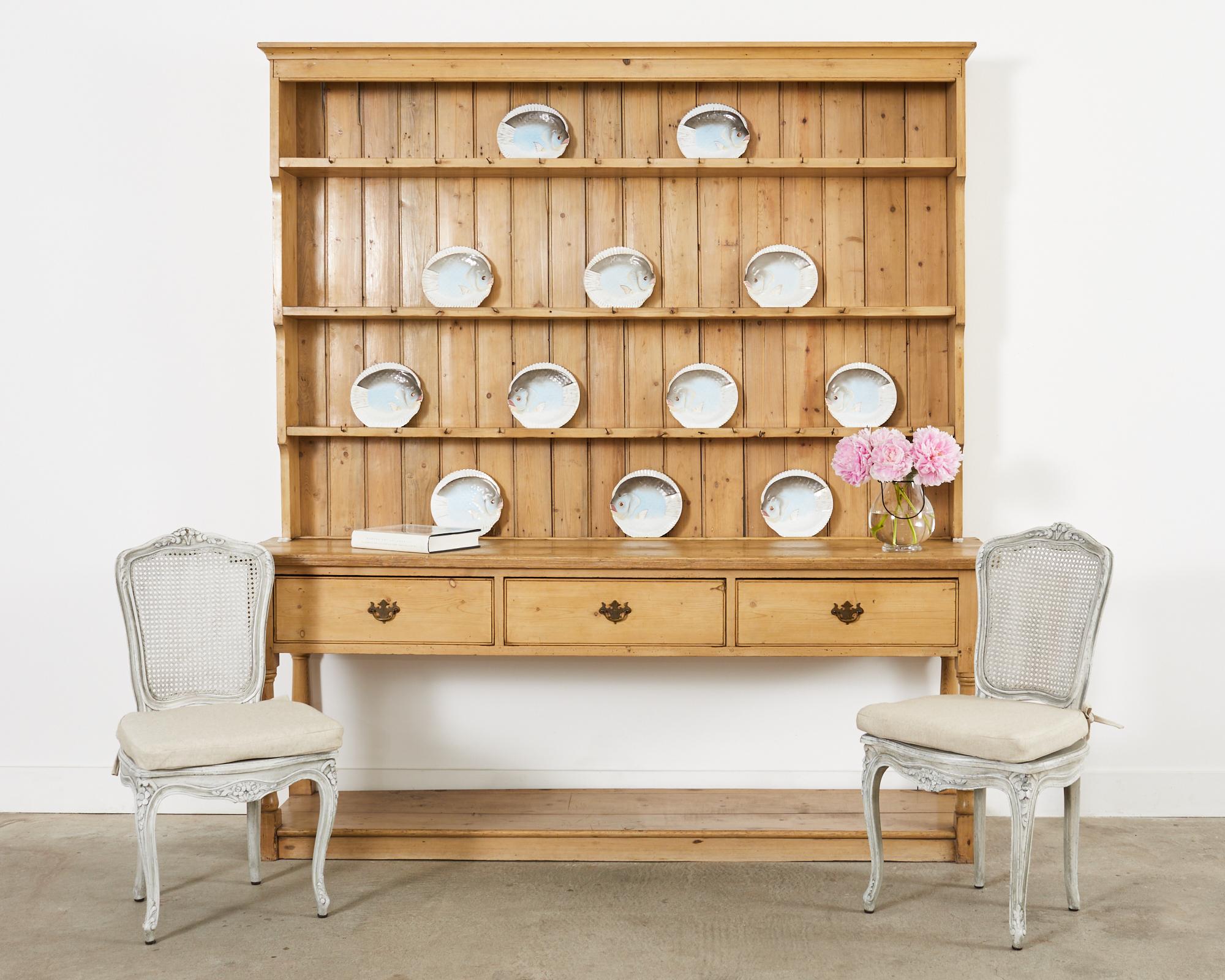 Rustic country English welsh cupboard dresser with pot rack storage crafted from pine. The grand cupboard rack measures 7 feet wide with three plate shelves surmounted by a cornice molding. The dresser is fronted by three large storage drawers with