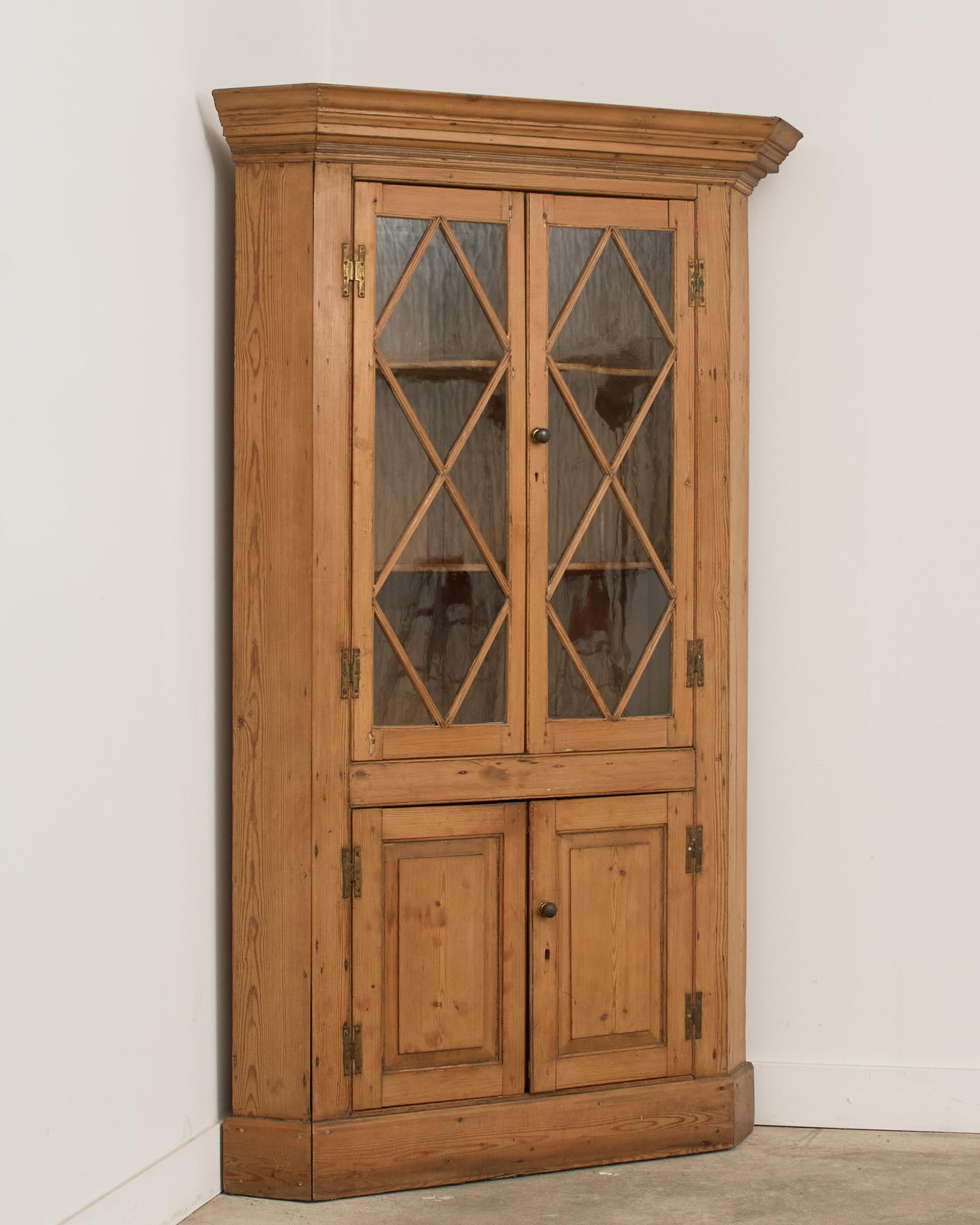 19th century gorgeous country English provincial cupboard corner cabinet or bookcase crafted from pine. The cabinet features a pair of glazed doors above a pair of paneled doors. The top has a cornice molding that angles back on the ends. The pine