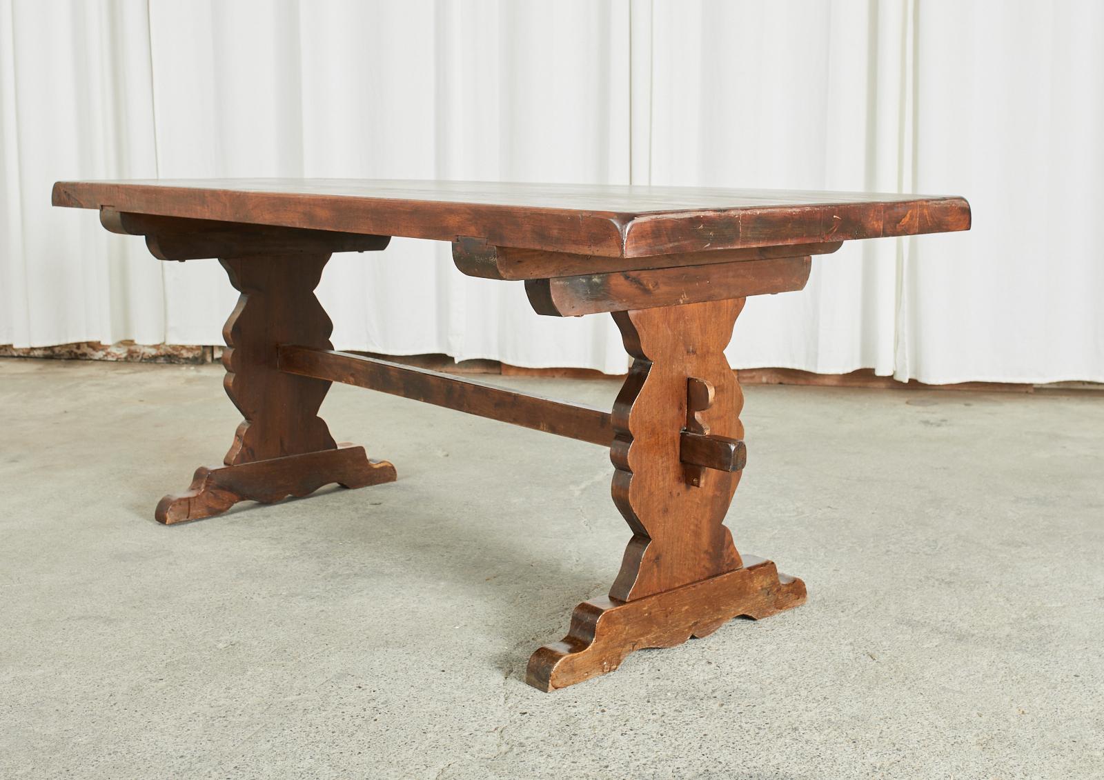 Rustic country French farmhouse dining table featuring a trestle style base. Crafted from solid birch with a 2 inch thick plank top that showcases the radiant woodgrains. Supported by baroque style thick shaped legs ending with shoe feet. The legs