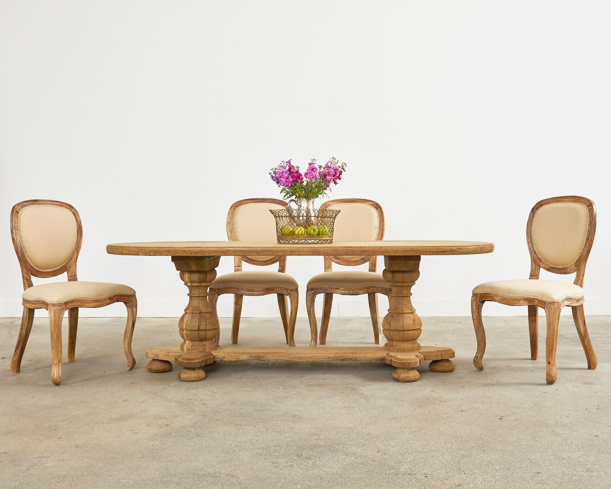 Rustic country French farmhouse trestle dining table featuring a bleached oak finish. The table has a large 2 inch thick plank top in an oval shape with a subtle ogee edge. The trestle style base affords ample legroom measuring 28 inches from the