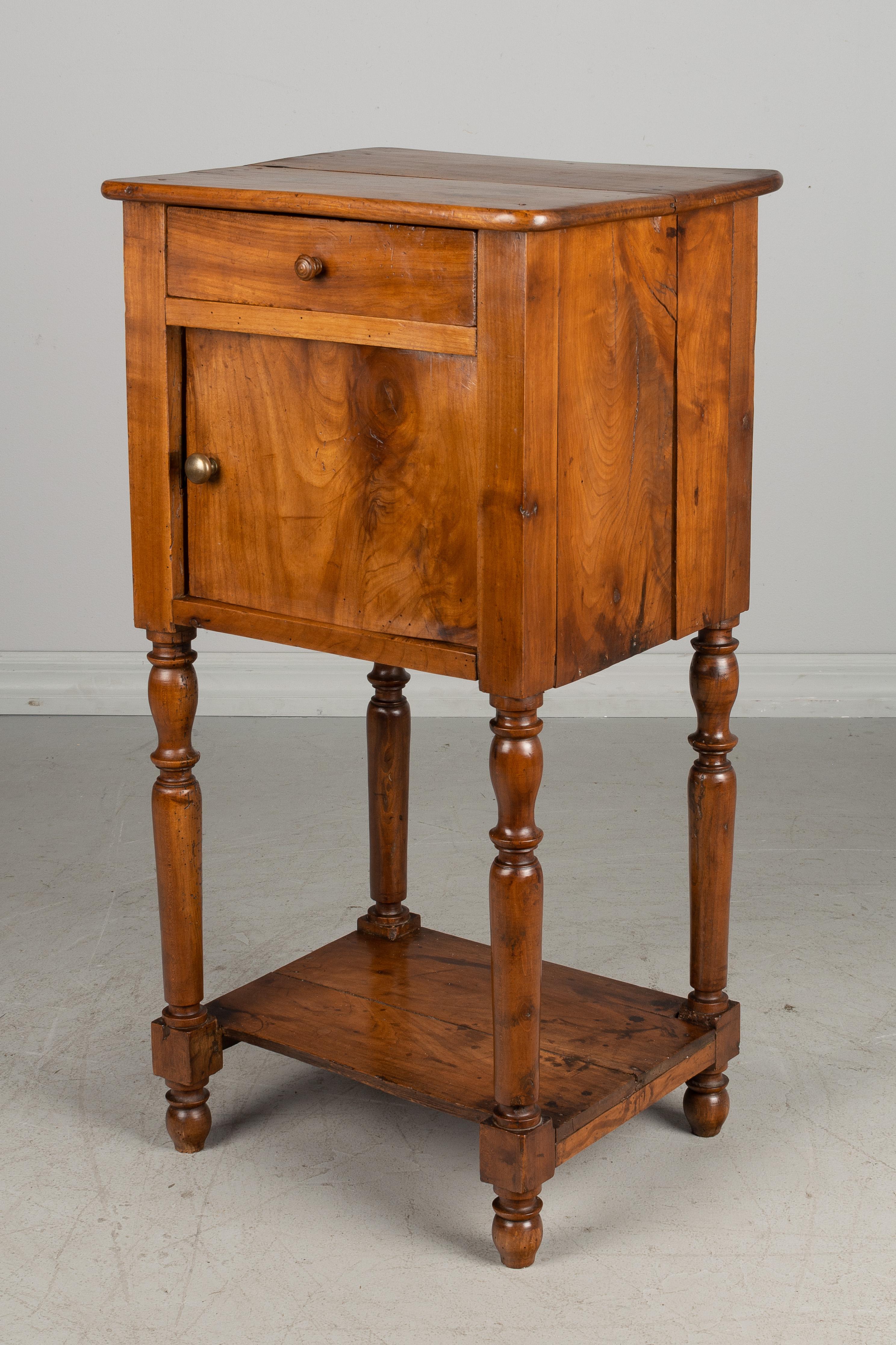 An early 20th century Country French tall side table or stand made of solid cherry with turned legs and lower shelf. Shallow drawer above a cabinet door opening to a fabric lined interior. Good restored condition.
