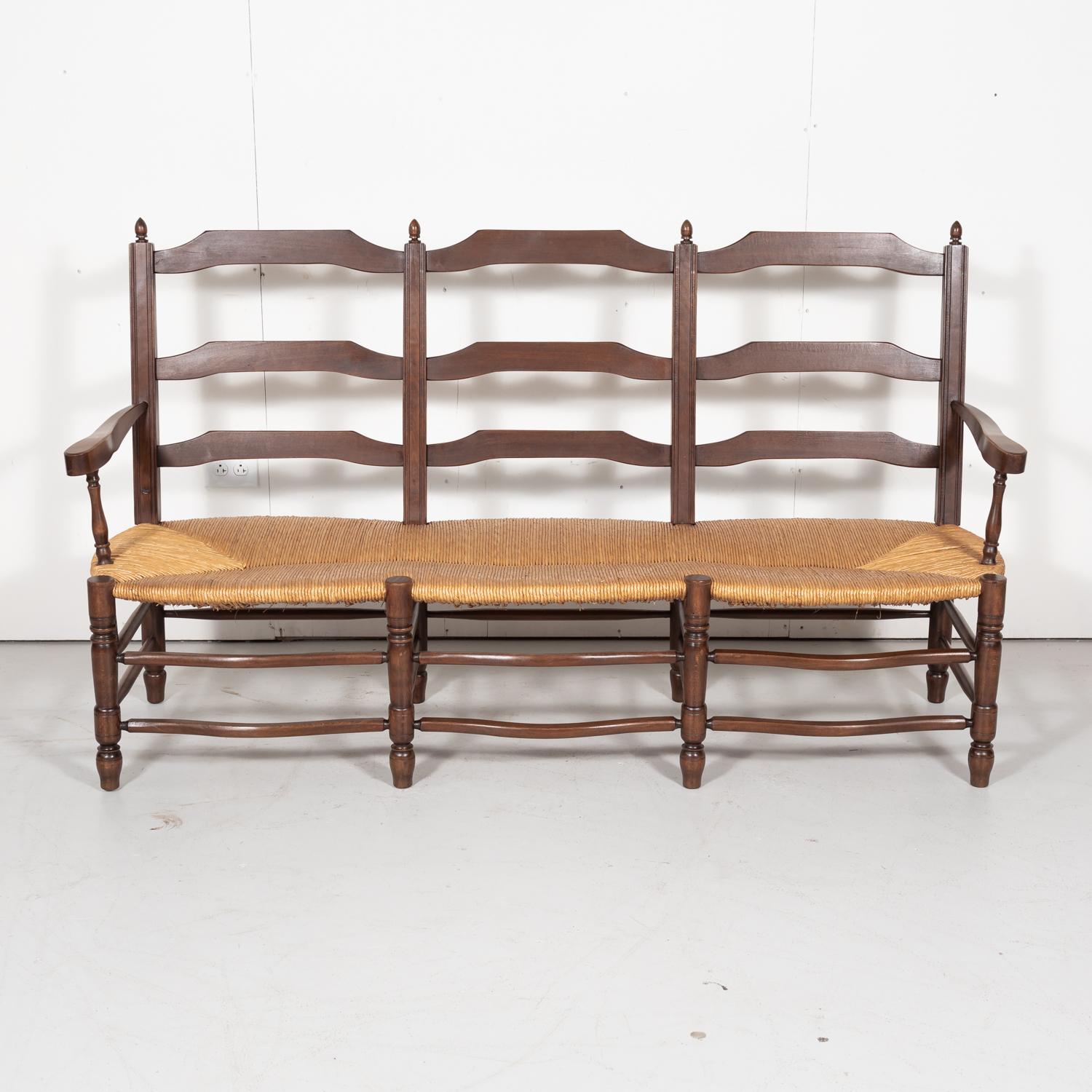 Country French settee or radassier in walnut, circa 1930s. This double arm Normandy bench seats three and features a handwoven rush seat and ladder-back. A wonderful rustic look that captures the charm and quaintness of rural France. This bench