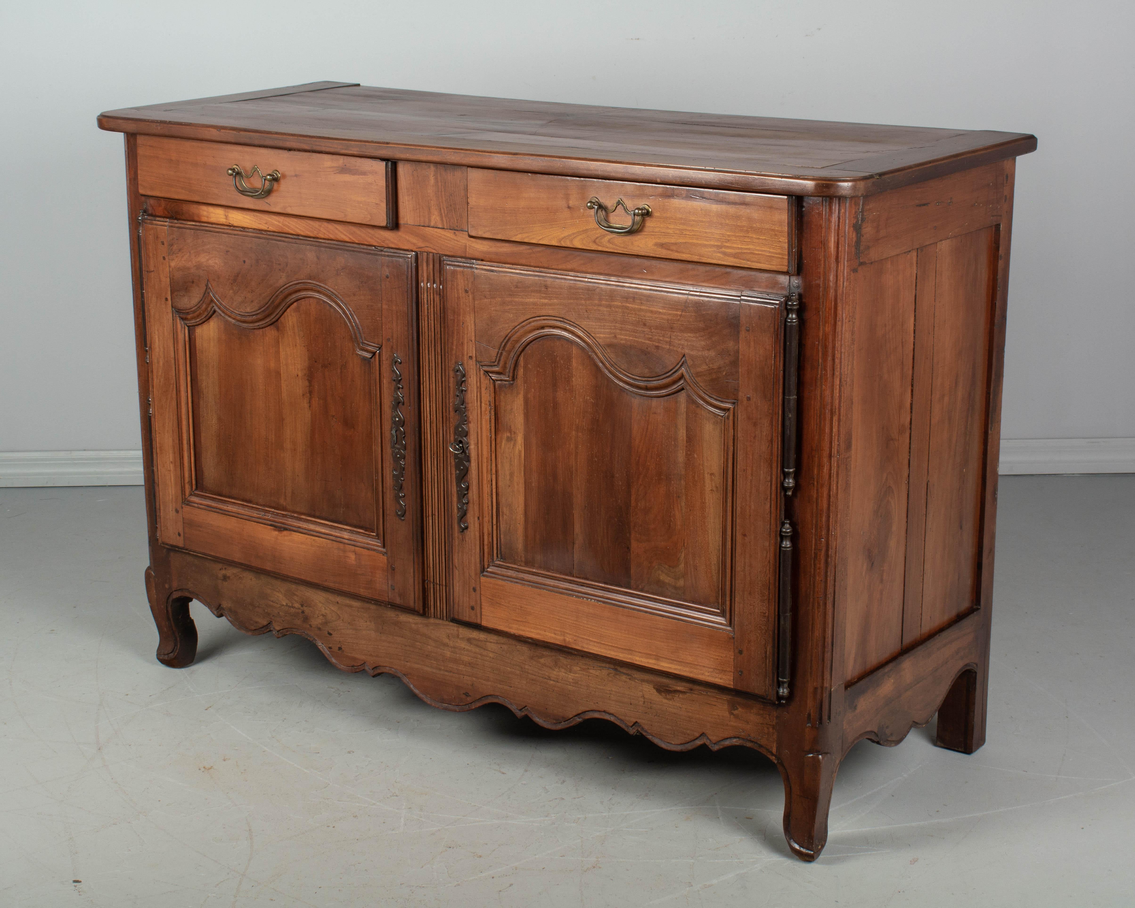A 19th century Louis XV style country French buffet from Normandy made of solid cherrywood. Simple hand carved panel doors and scalloped apron. Interior provides ample storage, opening to a single shelf. Working lock and key. Two dovetailed drawers