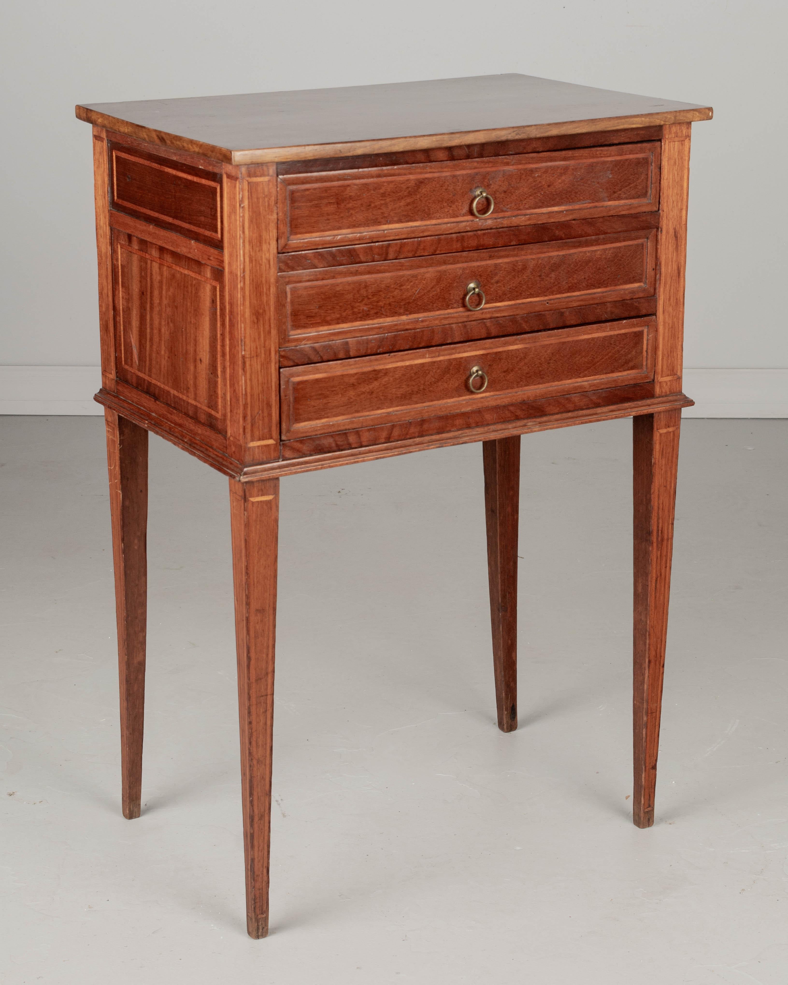 A Country French side table, handcrafted of solid mahogany and finished on all four sides. Three dovetailed drawers with small brass ring pulls. Sturdy slender tapered legs. Pegged construction. Waxed patina. Good proportions for use as a nightstand