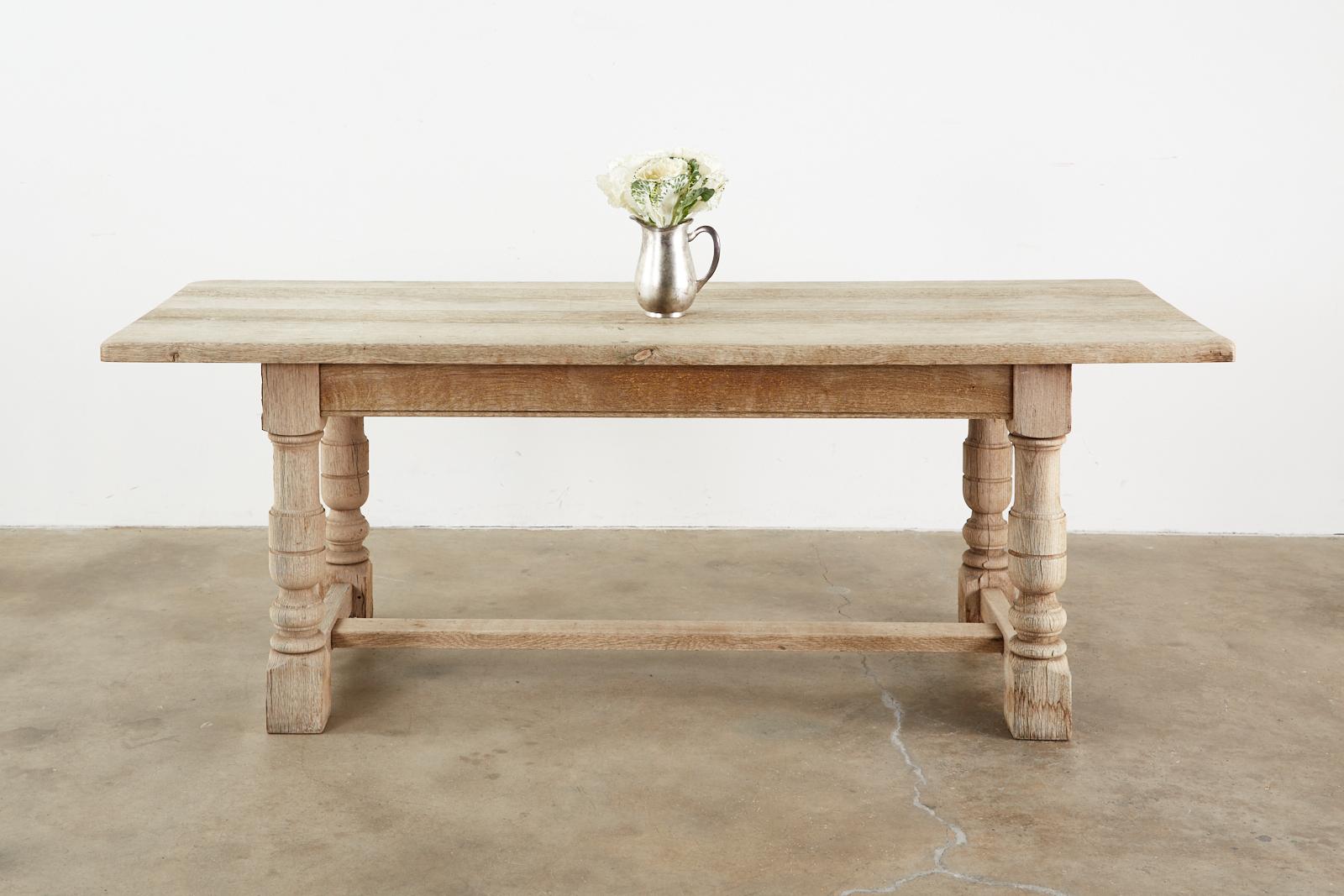 Rustic country French farmhouse dining table featuring a bleached oak distressed finish with an aged patina. Made in a grand trestle style with 1.5 inch thick plank top and thick, chunky turned legs ending with block feet. The legs are conjoined by