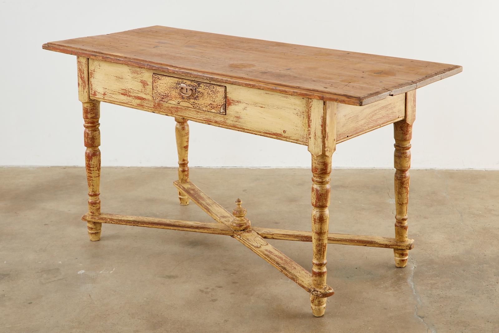 Charming country French pine farmhouse work table or console table featuring a distressed painted finish and patina. The base is fronted by a single storage drawer and supported by round turned legs conjoined by X-form cross stretchers having a