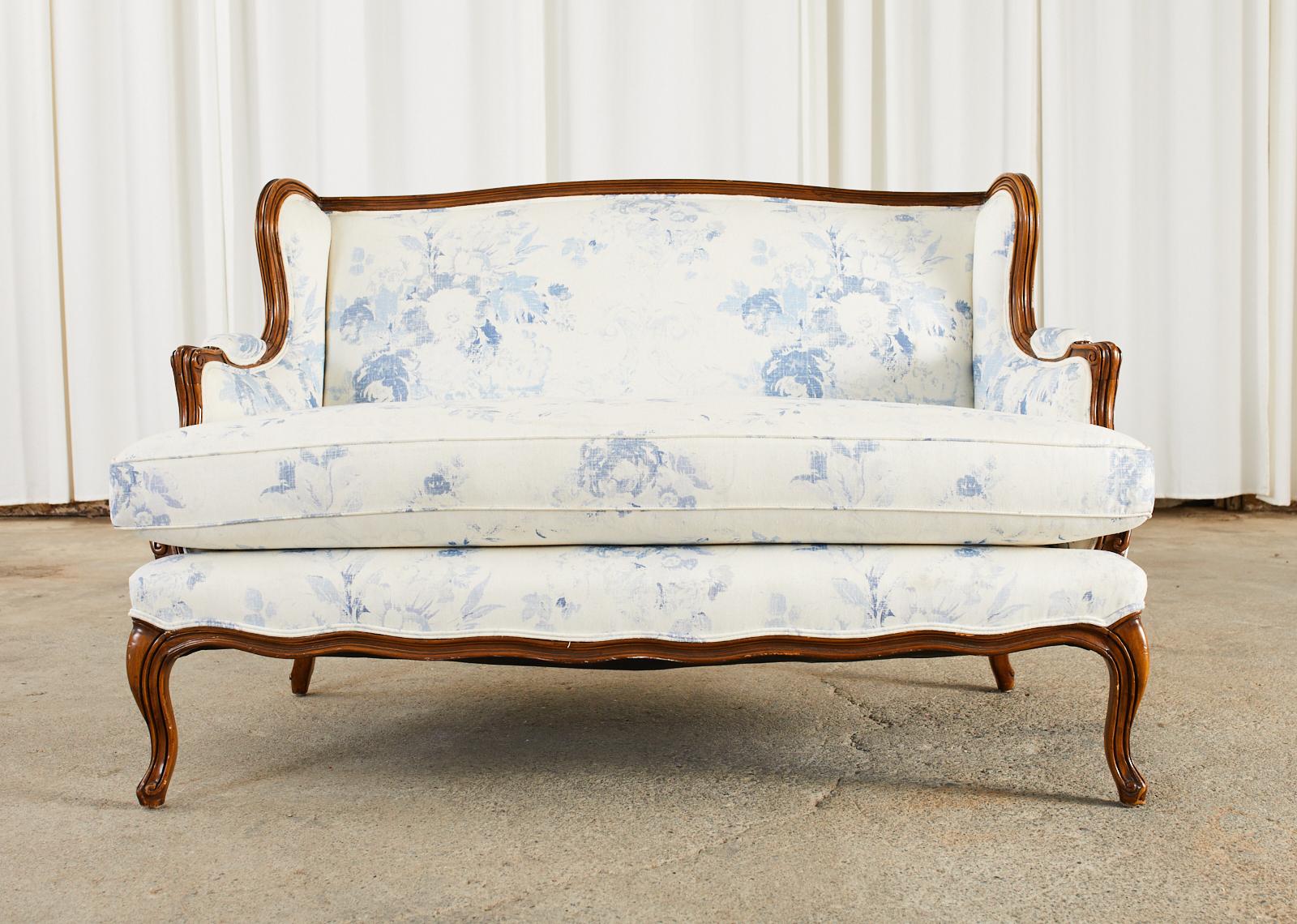 Stunning country French provincial wingback settee crafted from walnut featuring a dramatic blue and white upholstery. The settee has a gracefully curved frame with padded arms and a generous seat having a thick fitted seat cushion. The seat has a