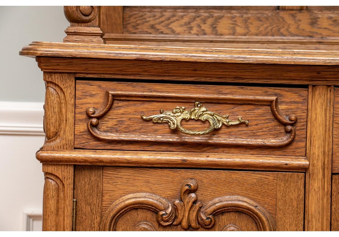 A very well made Country French Style cabinet with several finely crafted details. A tall two-part cabinet with shaped carved crest with open cartouche. The top cabinet with two side doors with carved floral scrolls and shelves, the center door with