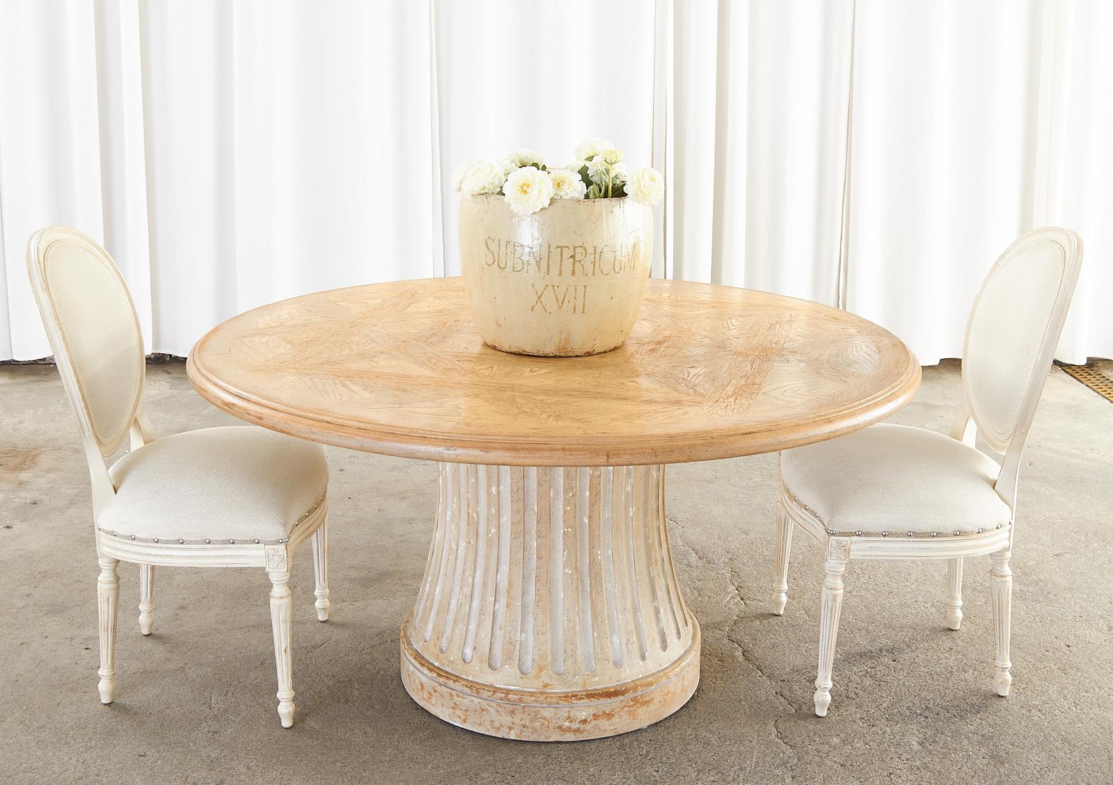 Distinctive round pedestal dining table featuring a round blonde oak top with parquetry inlay made in the country French style. The top is supported by a neoclassical style fluted column base with a distressed cerused finish on the waisted form. The