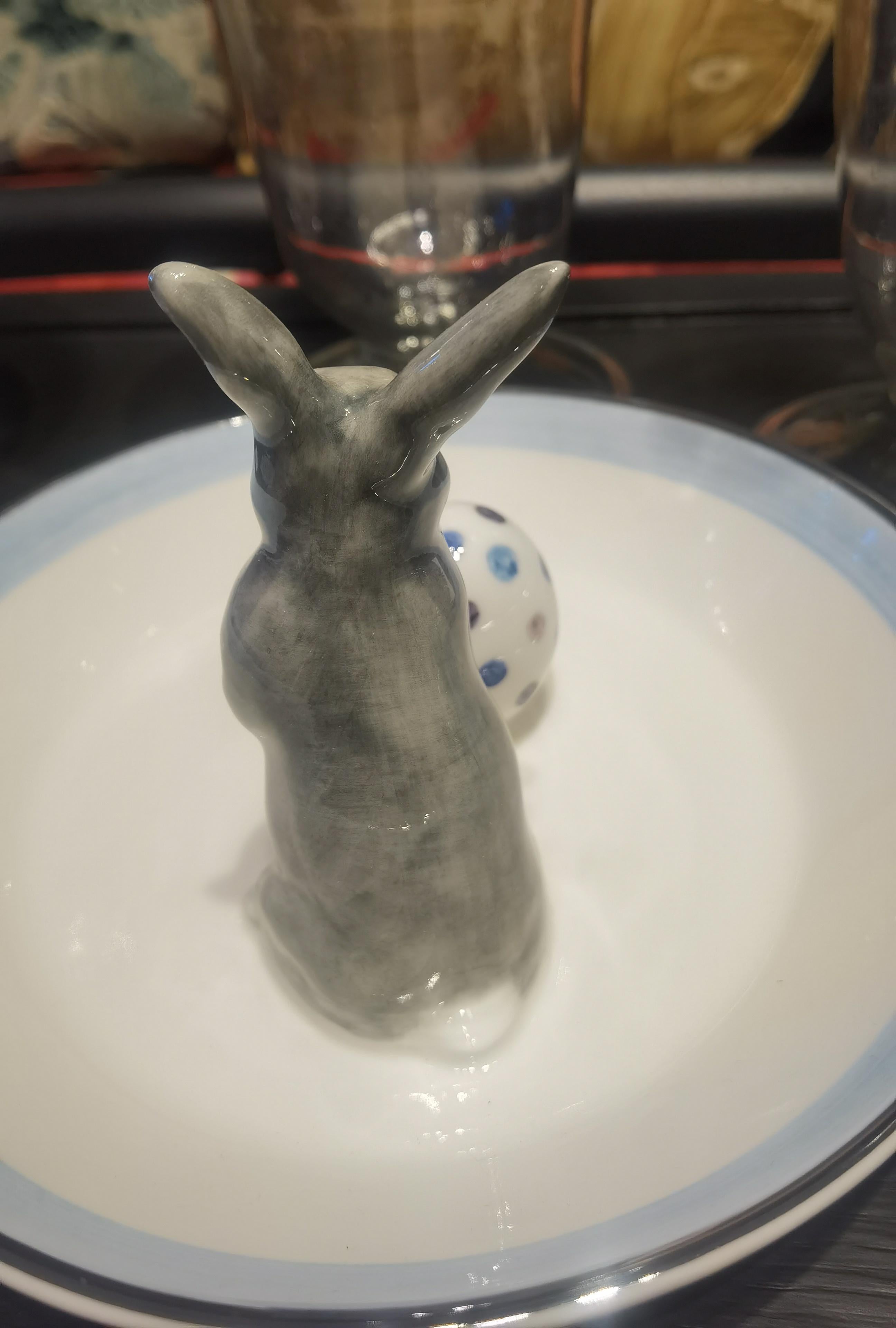 Cuenco de porcelana totalmente artesanal con una figura de liebre pintada naturalista en gris y blanco con un huevo. El conejo de Pascua está sentado junto a un huevo pintado a mano para decorar frutos secos o dulces alrededor. Ribeteado con una