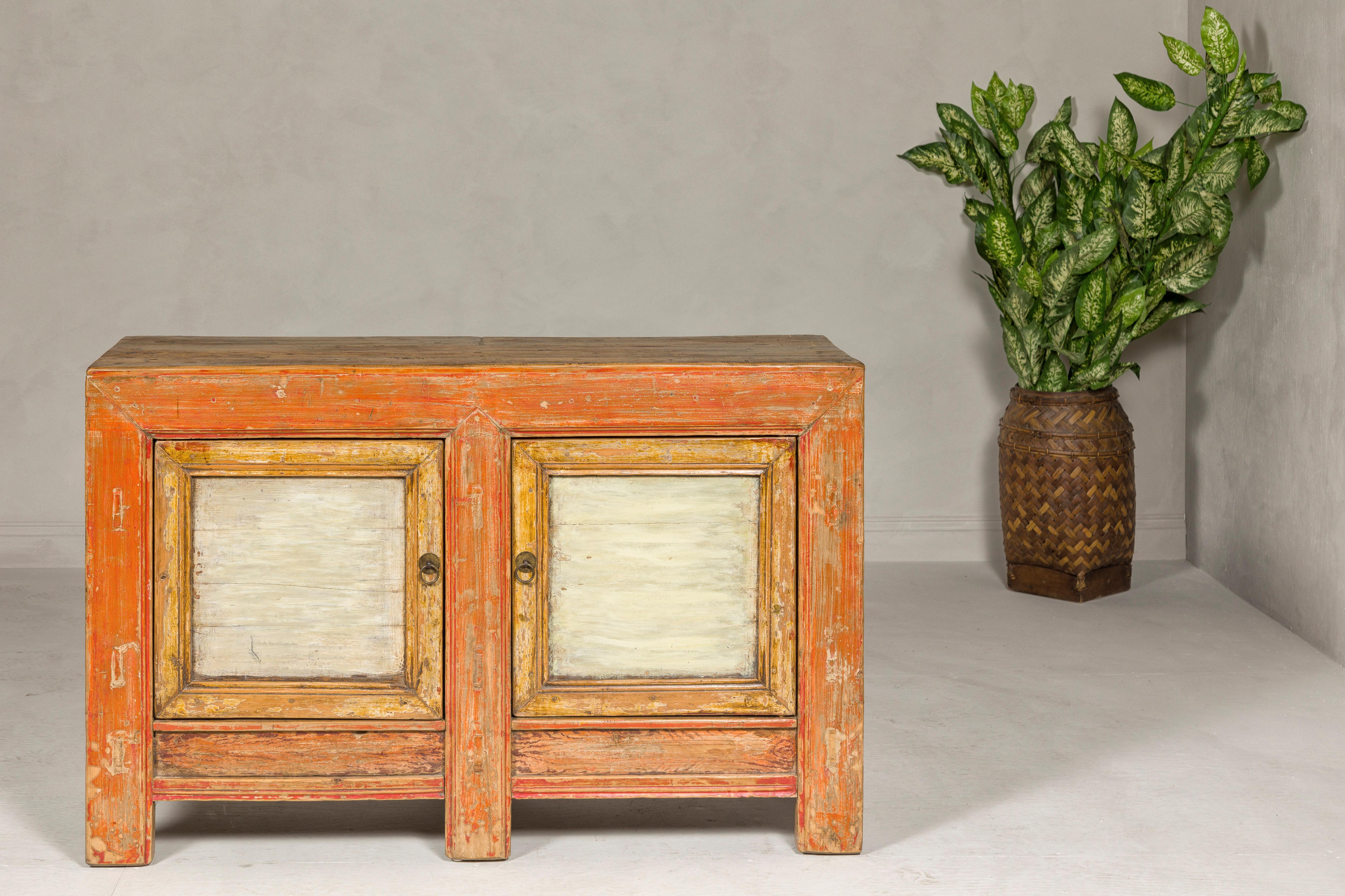 A Country style two-door buffet from the Midcentury period with distressed orange and off-white hand-painted color. Experience the charm and warmth of this Country style two-door buffet from the Midcentury period, a piece that beautifully showcases