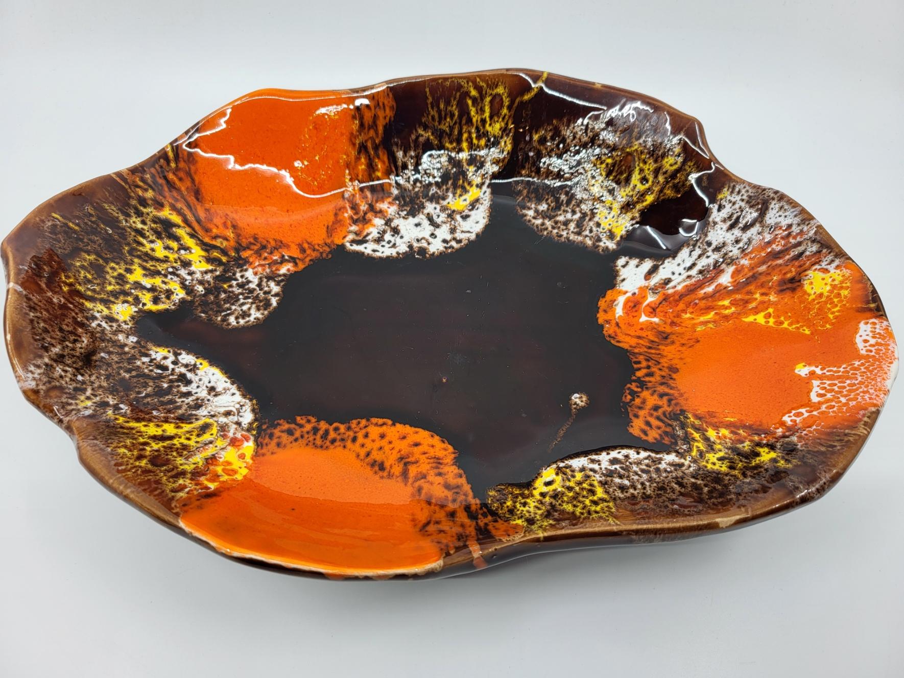 Cette magnifique coupe tripode de Vallauris, en céramique émaillée française, présente un design unique et captivant. Sa couleur de base marron, harmonieusement enrichie de glaçures orange et blanche, lui confère une allure chaleureuse et