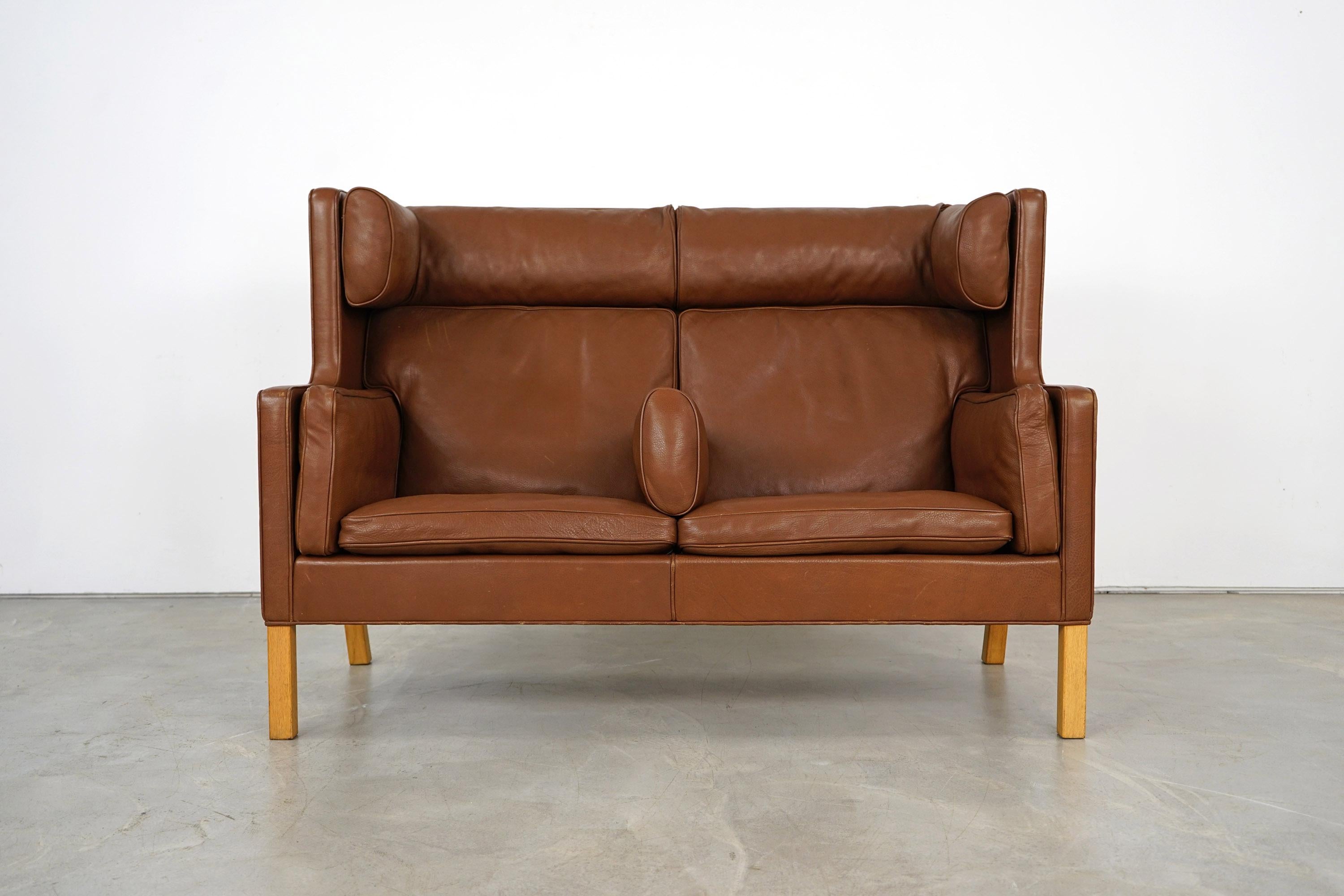 Børge Mogensen's Coupe 2-seat sofa was produced by the manufacturer Fredericia in Denmark. The piece is from the 1970s. The high-backed piece of furniture stands on legs made of light wood. The brown leather is in a very good vintage condition and