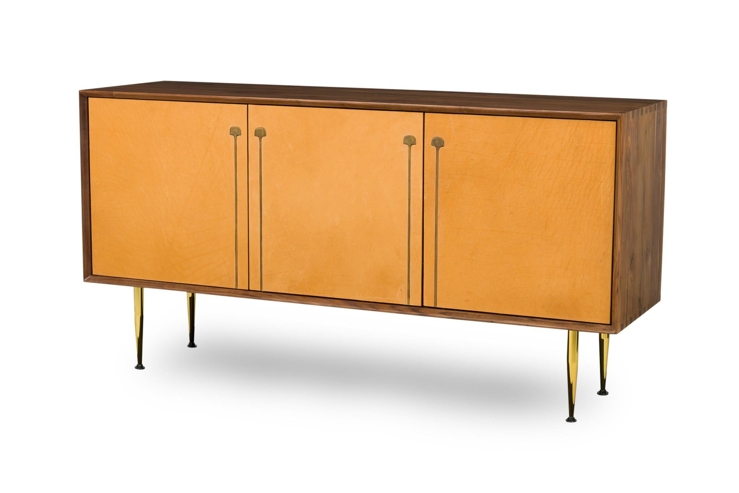 Contemporary / Mid-Century inspired credenza constructed of sustainably harvested FSC-certified wood and features elegant bronze inlaid leather upholstered doors which open up to ample storage, sitting atop tapered brass legs. 
 

