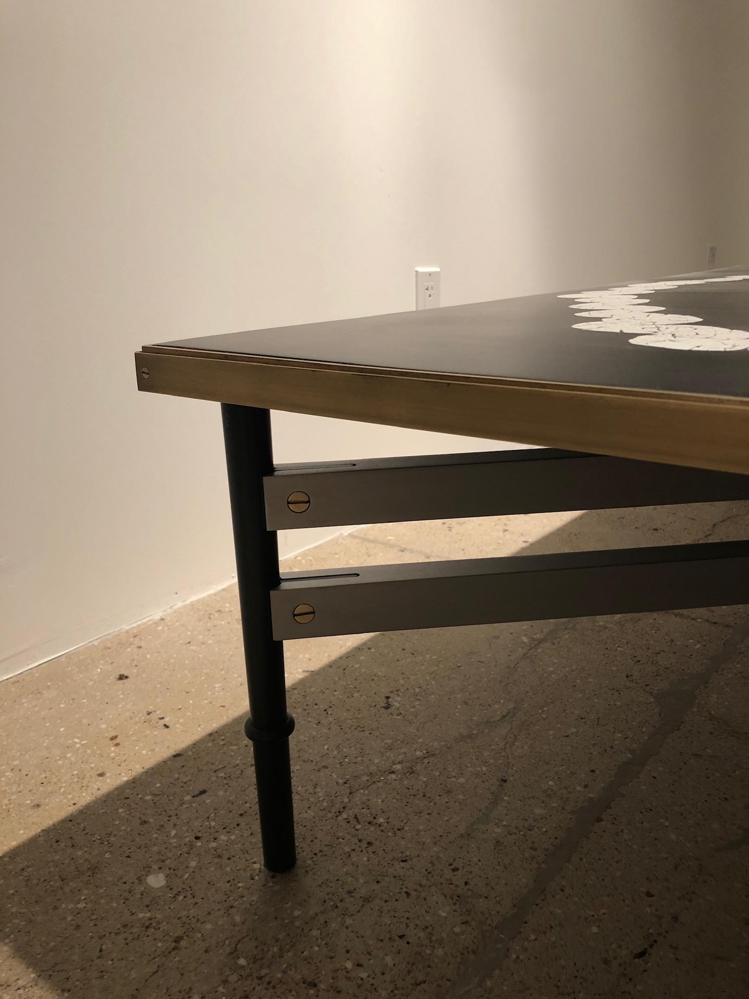 This coffee table features a hand-applied ring of cracked eggshell trapped in resin and lacquer, finished with a satin brass trim around the edges. The base is structured by a combination of blackened steel legs with satin aluminum stretchers.