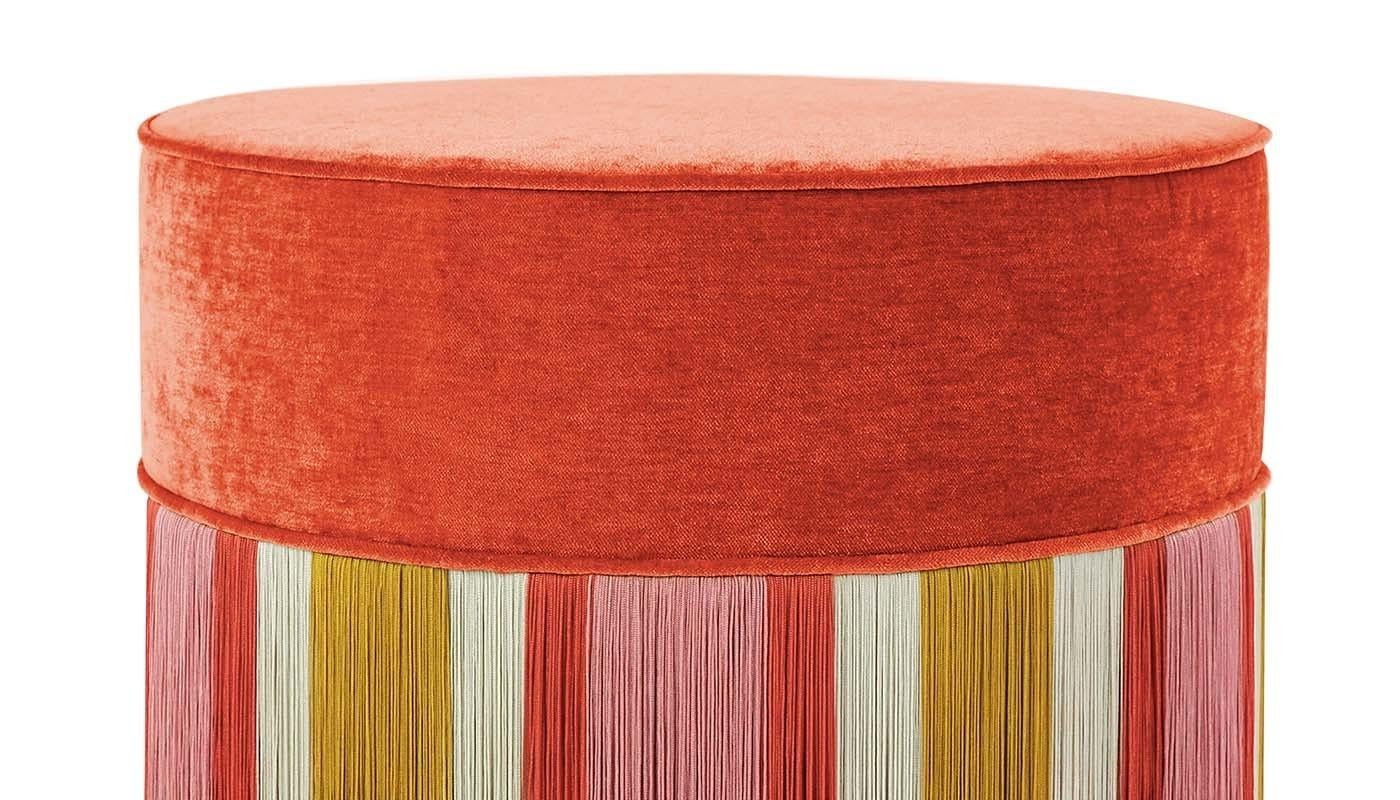 This Art Deco-inspired ottoman is entirely upholstered in velvet made of acetate (53%), polyester (42%), and viscose (5%). It stands out for its bright colors and sophisticated geometric pattern: the cover comprises a solid orange top and fringes in