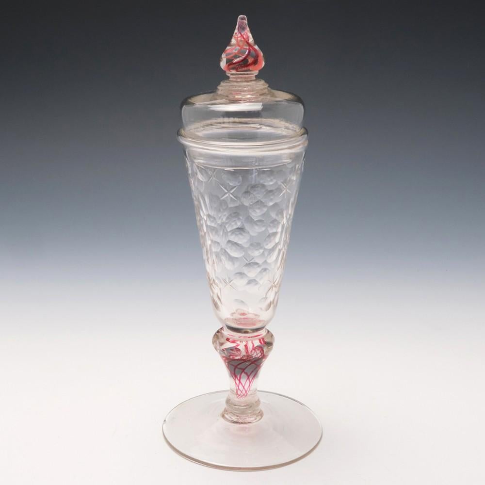 Covered Wine Goblet With Colour Twist Stem, c1730

The tall funnel shaped bowl is  cut with three flowers and then bordered with lens and star cuts.  The stem has a five thread  ruby red wist which is mirrored in the finial.  The cover sits