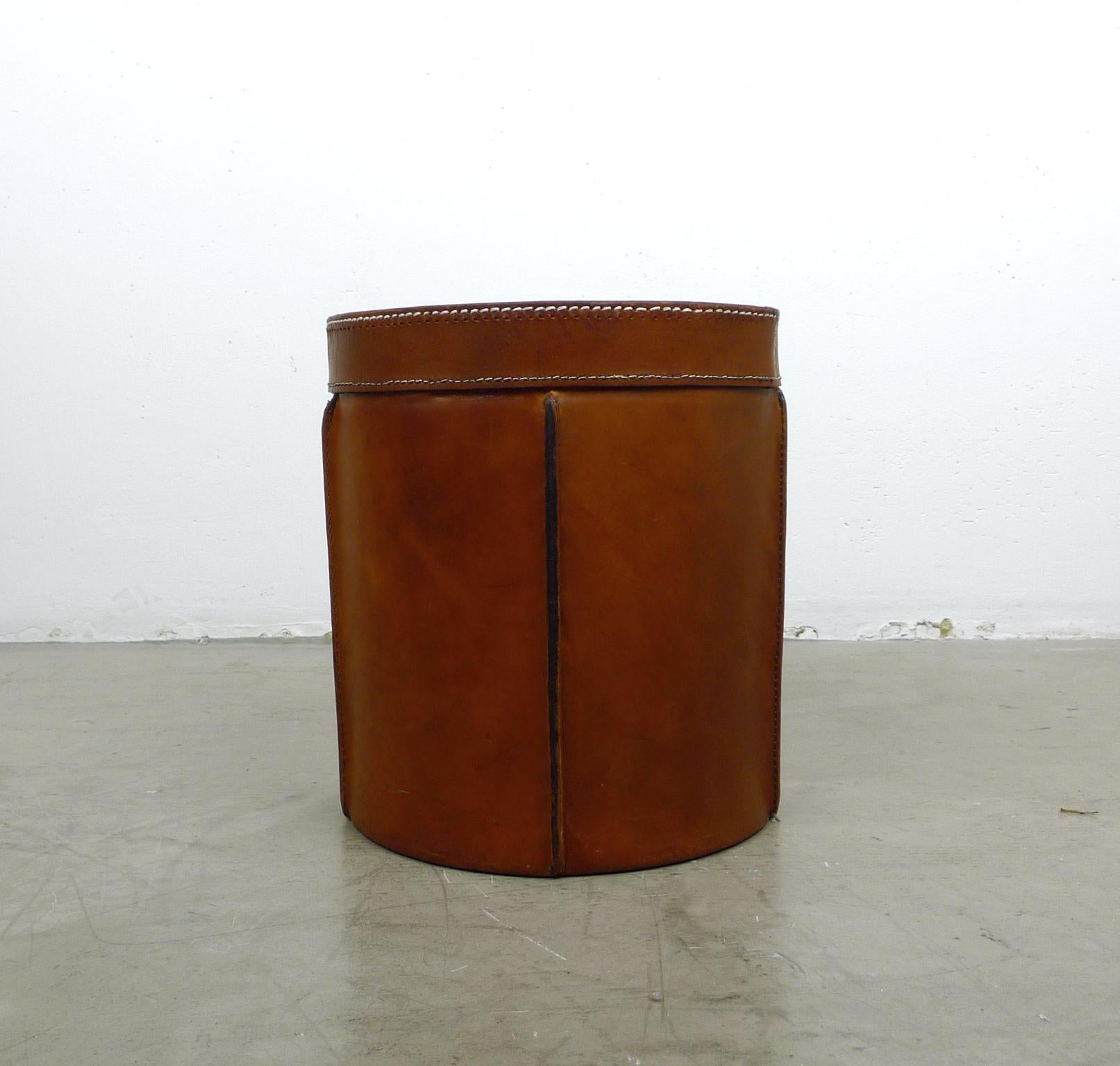 Round paper basket made of thick cognac-colored cowhide from the 1960s. The body is sewn in individual leather elements with white yarn on vertical bars. The interior is lined with a lighter leather. It is in very good condition.