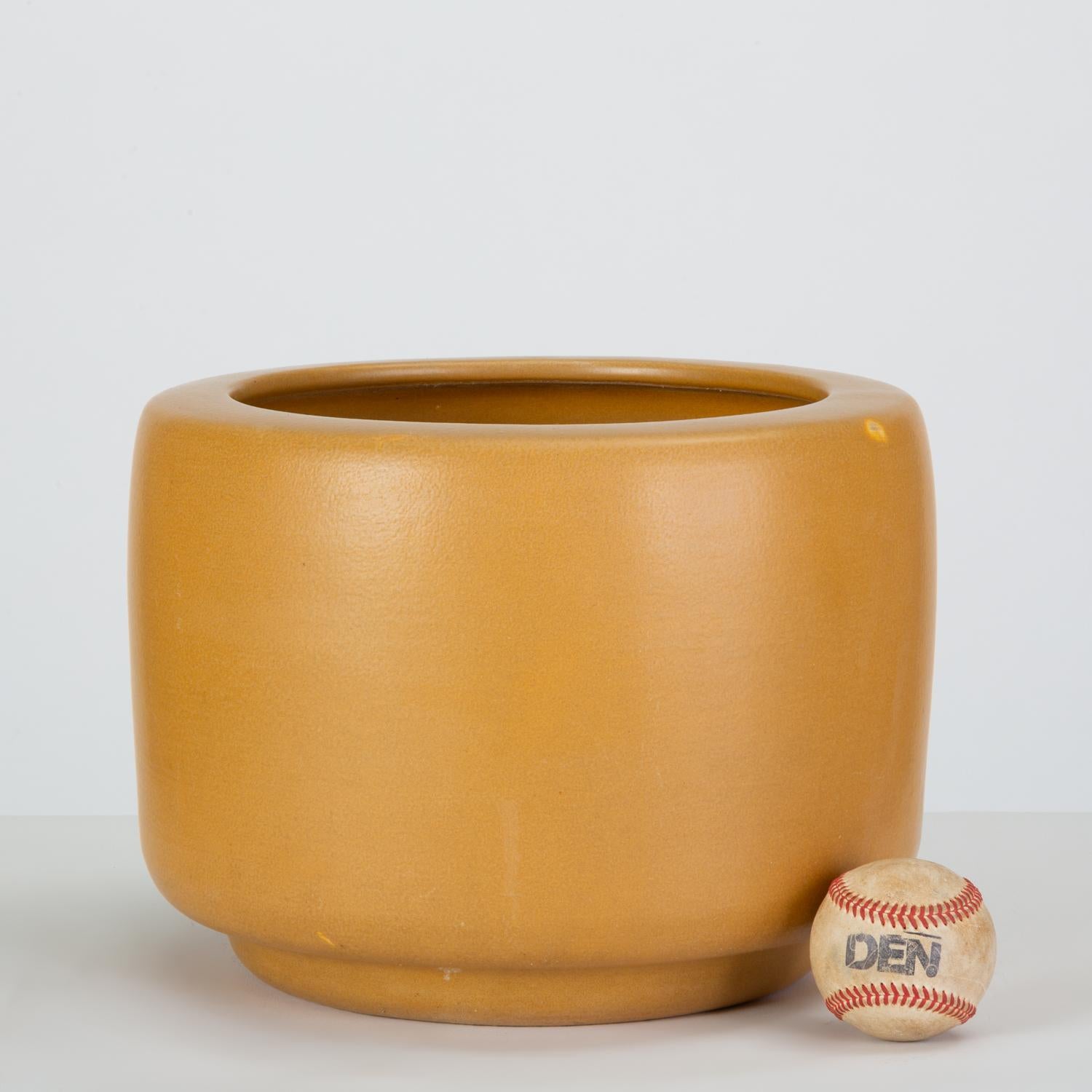 A mustard yellow bisque planter by John Follis for Architectural Pottery. This example has a drum shape with a rounded lip that curves in at the top of the piece. Planter retains partial stamp: “Architectural Pottery Made in USA.”

Condition: