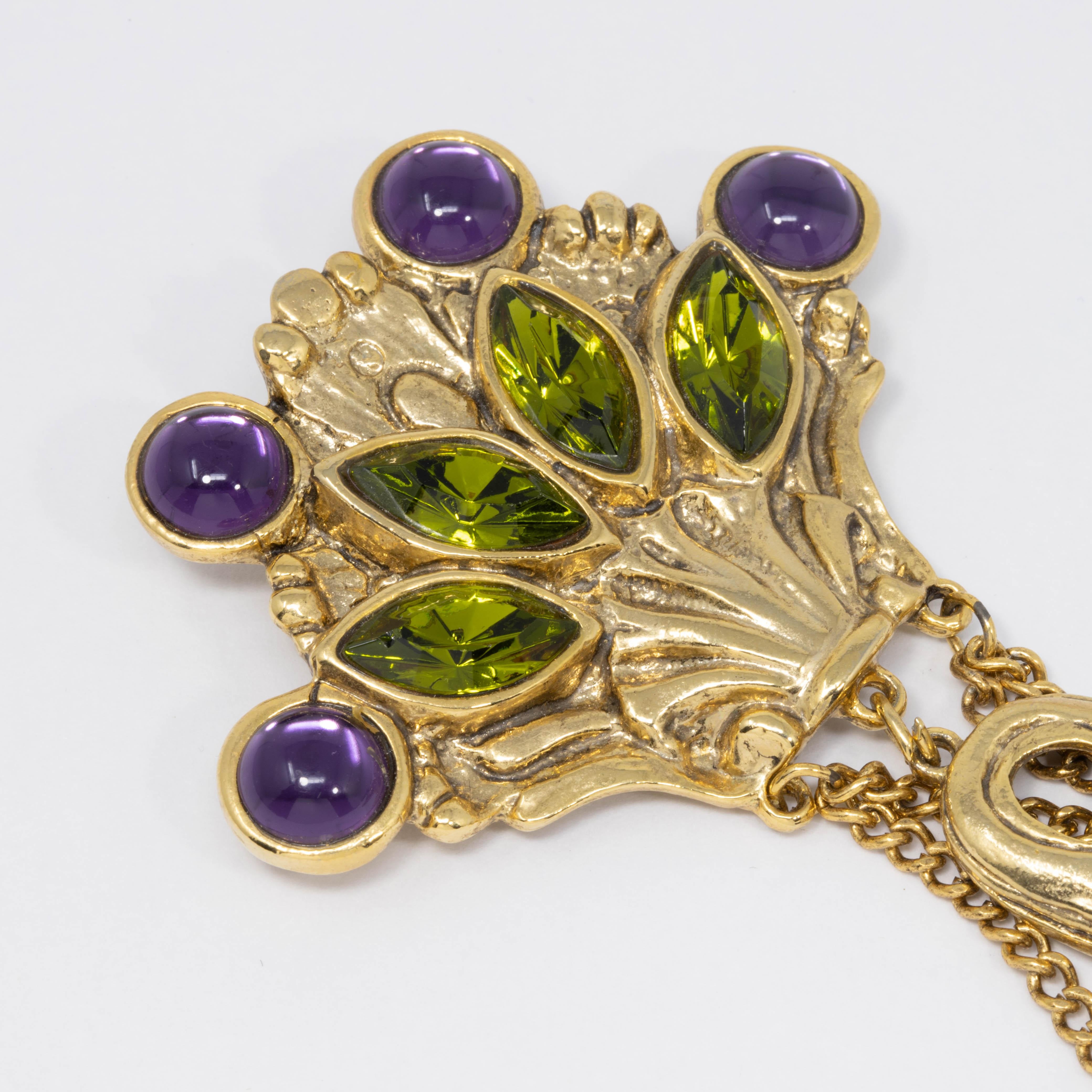 A dangling pin brooch by Craft. Influenced by Victorian and Art Nouveau style, this stylish accessory features a extravagant design with violet cabochons and green crystals, accented with a dangling chain and motif.

Signed CRAFT