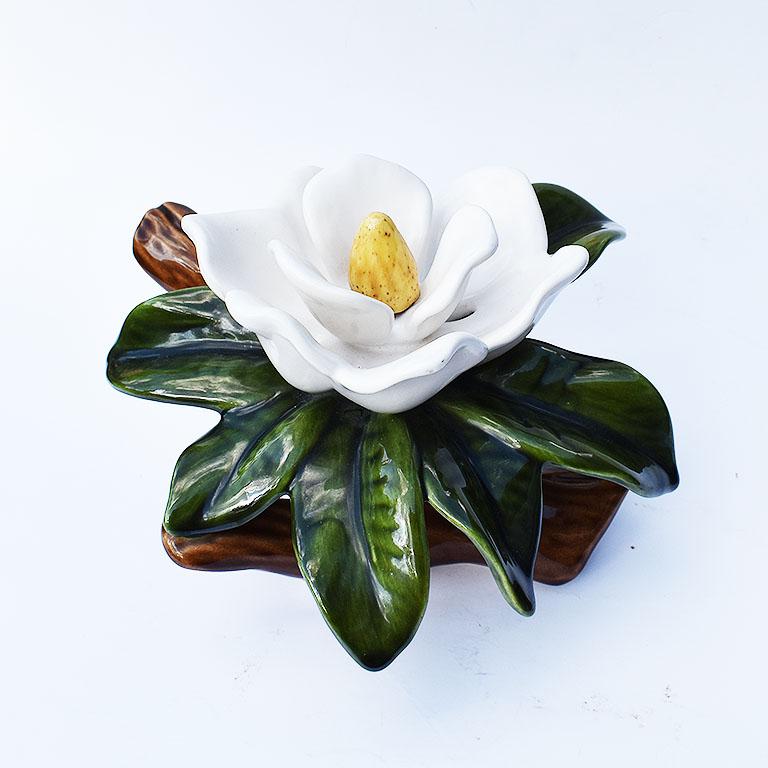 Ceramic Magnolia flower in green, white, yellow, and brown. This ceramic decorative piece shows a blossoming magnolia flower, with large full green leaves. The flower rests upon a brown ceramic branch. 

Specifications:
9