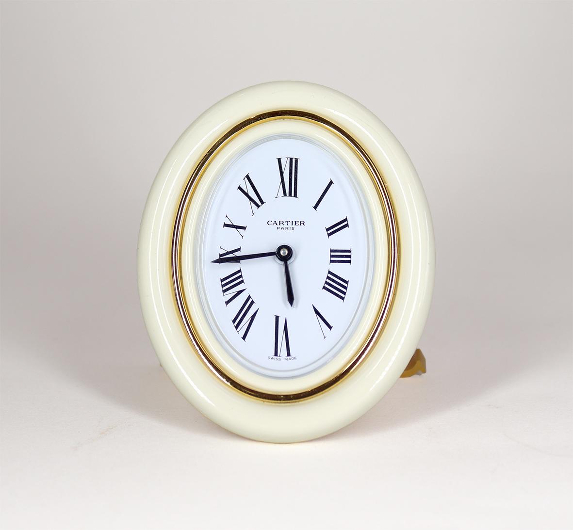 A Cartier Alarm Clock in an oval enamel and gold plated frame. The cream enamel and gilt frame is in perfect as new condition, with cabuchon jewelled setting nut, resting on a strut back with the distinctive Cartier ‘C’ forming the feet.

The oval