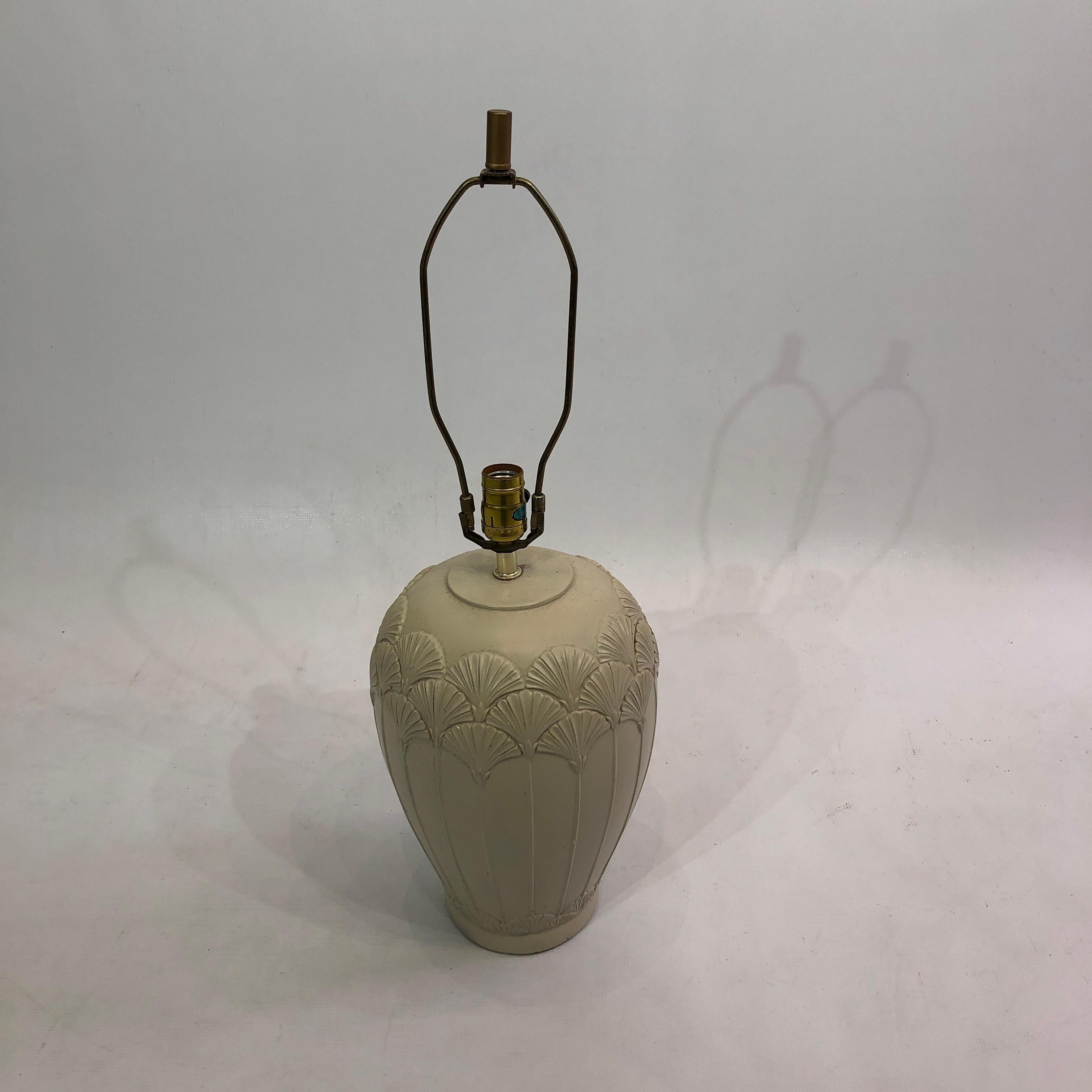 This Art Deco-inspired ceramic table lamp features an intricate shell pattern and comes in a faded cream colour. The lamp base is shaped like an urn, with a narrow bottom and heavier top. Protruding from this is an American style harp fitting for a